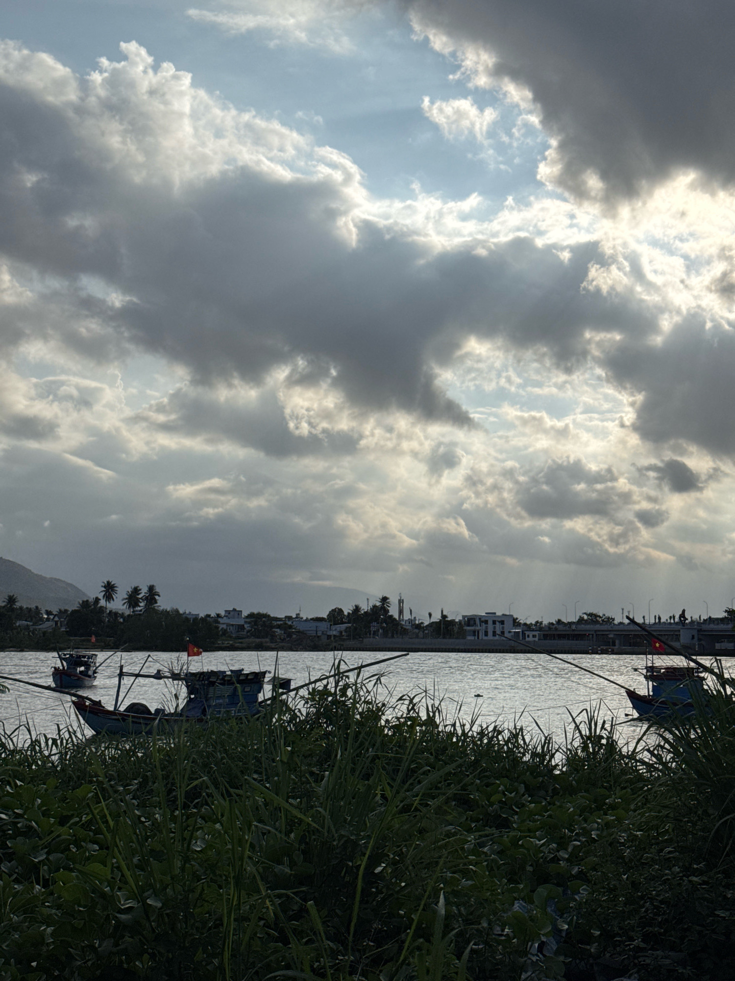 The image depicts a river scene in Nha Trang, Vietnam, during a cloudy day with shafts of sunlight breaking through. In the foreground, dense green vegetation lines the riverbank. The river itself is calm, with several fishing boats moored or navigating its waters. One prominent blue fishing boat in the mid-ground flies a Vietnamese flag. In the distance, a cityscape with buildings and palm trees is visible along the opposite bank, with a mountain range forming the backdrop. The sky is filled with dramatic, layered clouds, some dark and heavy, others illuminated by the sun. A bridge or elevated structure is discernible in the far distance.