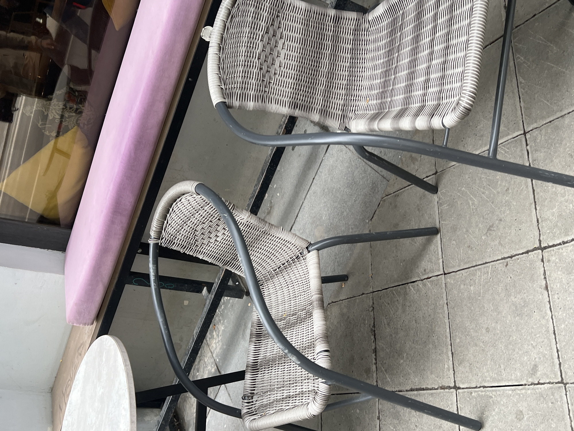 Two wicker chairs with metal frames sit on a paved patio in front of a window. A pink upholstered bench is visible behind the chairs. A circular table with a light-colored top is partially visible to the left. The scene appears to be an outdoor seating area for a cafe or restaurant. There are no people or animals visible in the image. The location is Yekaterinburg, Russia.