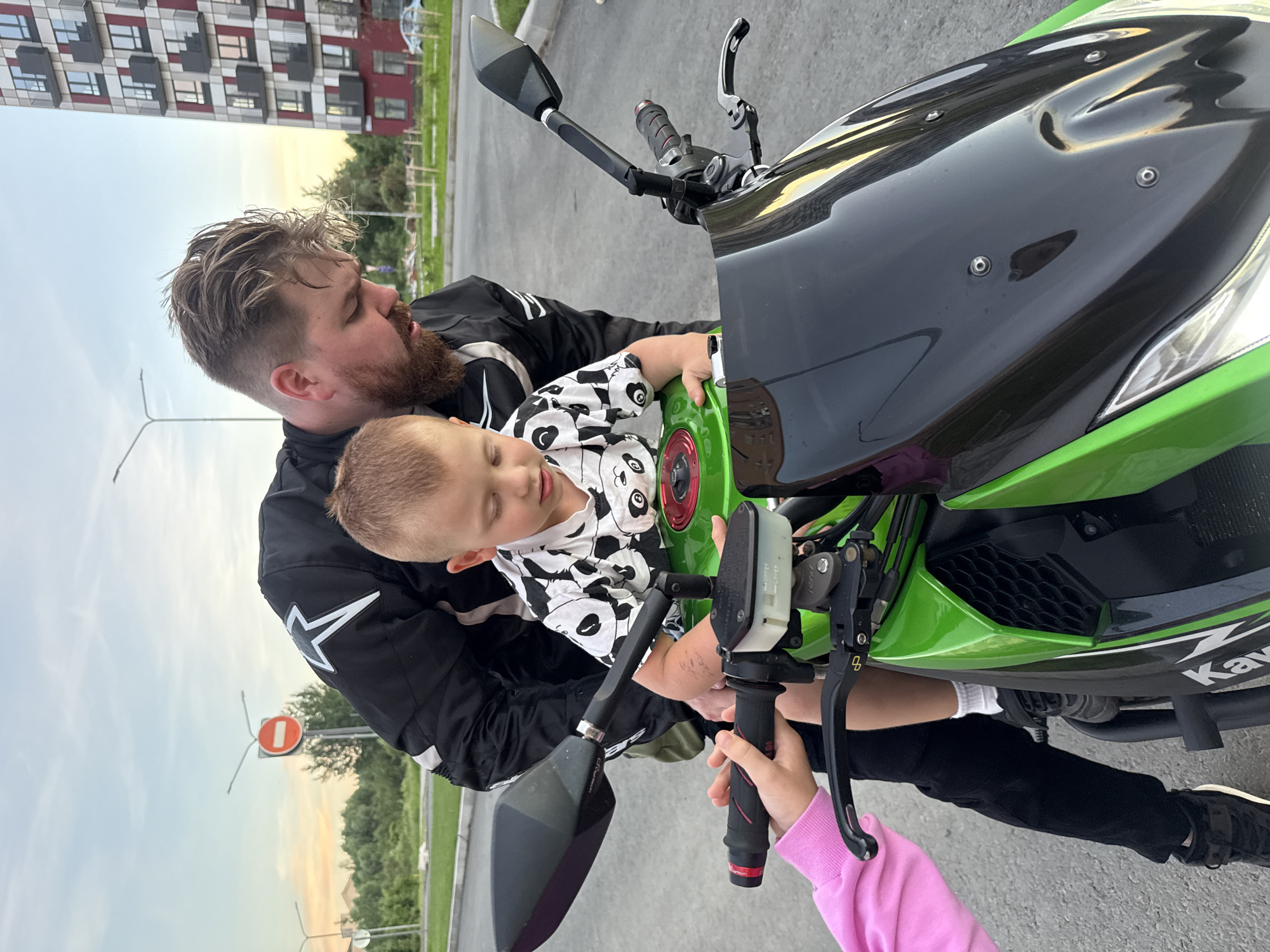 In the city of Veliky Novgorod, Russia, a man with a beard wearing a black motorcycle jacket, and a young boy in a black and white panda print shirt are sitting on a green motorcycle. The boy is in front, and the man is behind him, leaning forward. The boy's hands are on the gas tank, and the man's hands are on the handlebars. A child's pink-sleeved arm is visible on the left, also reaching towards the handlebars. The motorcycle is parked on an asphalt surface, likely a street or parking lot. In the background, there is a modern apartment building with red and white accents, trees, streetlights, and a road. The sky is partly cloudy with a sunset glow.