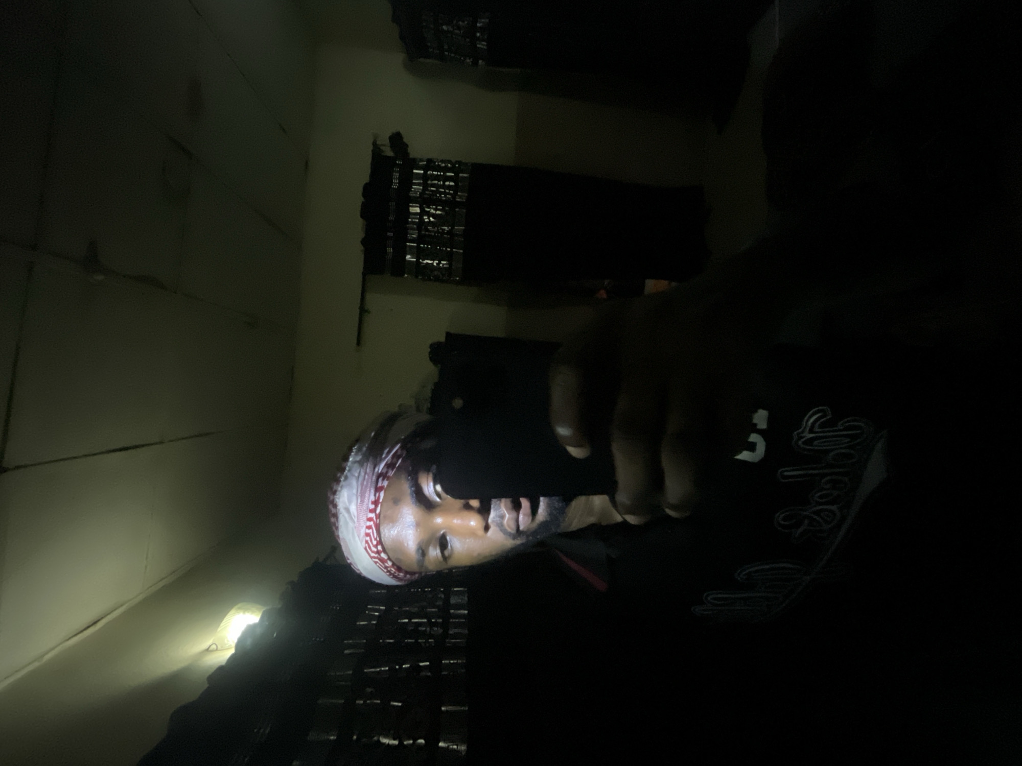 A man wearing a white and red patterned head covering and a black shirt with white writing is holding a black smartphone in front of his face, seemingly taking a selfie in a dimly lit room. The room has two dark window openings, possibly covered with something obscuring the view. The walls appear to be made of light-colored panels or boards. The exact location in Lokoja, Nigeria, cannot be determined from the provided image.
