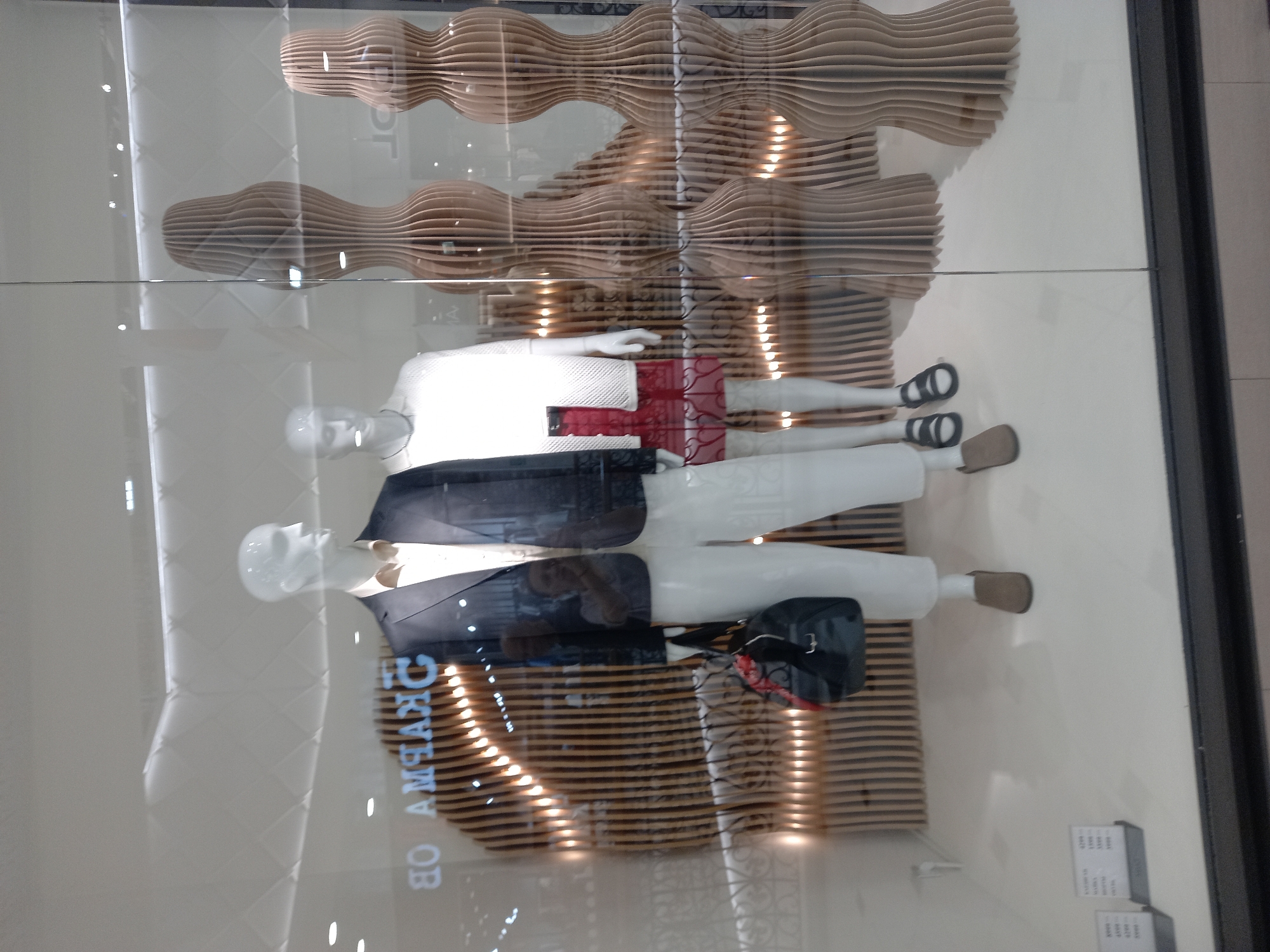 A store window display features two mannequins. The mannequin on the left is wearing a dark suit jacket over a light-colored collared shirt, white pants, and brown slip-on shoes. It is also holding a black handbag with red accents. The mannequin on the right is wearing a white open-knit cardigan over a white top and a red skirt. It has black sandals. The background of the display consists of wooden, wavy decorative elements and a sign with Cyrillic text that reads "OB AMHAHC." The display is located in Yekaterinburg, Russia.