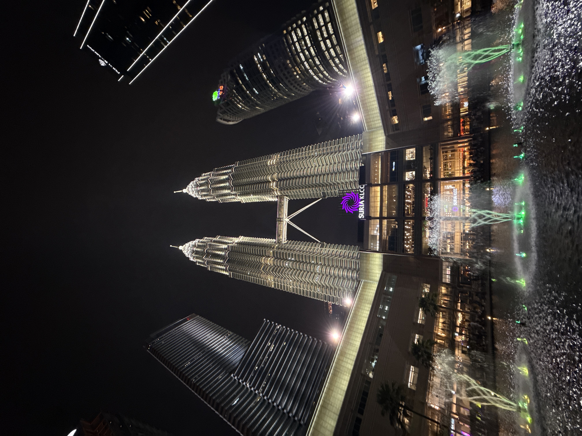 The image shows a night view of the Petronas Twin Towers in Kuala Lumpur, Malaysia. The towers are illuminated, with other surrounding skyscrapers also visible. A fountain with green-lit water features is in the foreground, reflecting the buildings.
