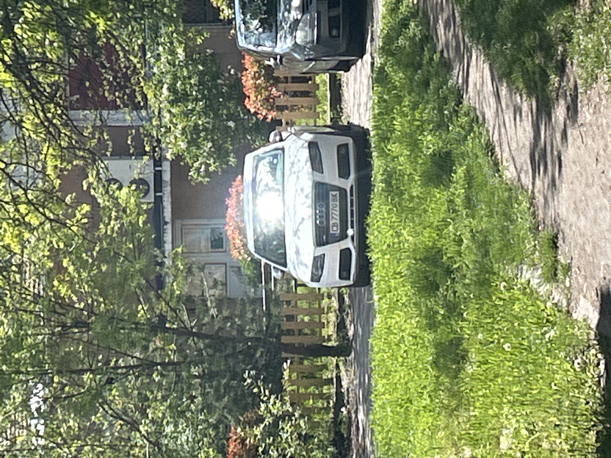 The image shows a street scene in Sofia, Bulgaria. A white Audi SUV with license plate "CB 7770 BK" is parked on the side of the road. A dark-colored car is parked behind it. A sidewalk runs along the right side of the image. A building is visible behind the cars, partially obscured by trees. A fence runs parallel to the sidewalk. The scene is bathed in sunlight.