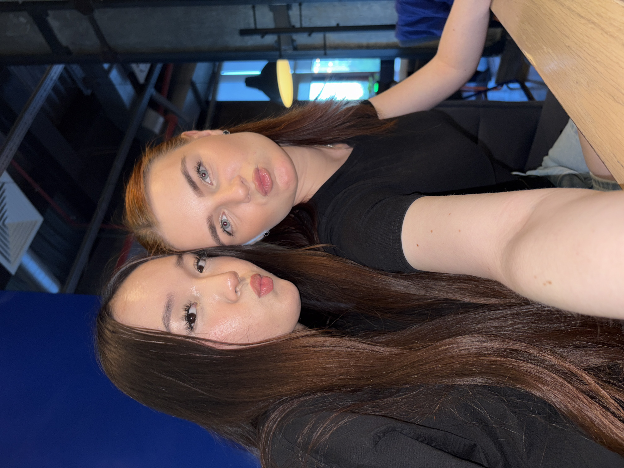 This image depicts two young women posing for a selfie indoors, presumed to be in the city of Sinyavino, Russia.
The woman on the left has long, straight, dark brown hair with lighter reddish-brown highlights towards the ends, parted in the middle. She has light skin and appears to be of East Asian descent, with visible makeup including long eyelashes and lip gloss. Her lips are puckered. She is wearing a black long-sleeved top.
The woman on the right has medium-length reddish-brown hair, pulled back from her face, with light blue-grey eyes and light skin. She is also making a similar puckered lip expression. She wears a black short-sleeved t-shirt, a small black stud earring in her left ear, and a white wireless earbud in her left ear. Part of her light-colored shorts is visible below a table.
Both individuals are looking directly towards the camera. The foreground features a light-colored wooden table. The background on the left consists of a solid dark blue wall. The background on the right is a modern indoor space with a dark ceiling revealing exposed pipes and ventilation, dark vertical structural elements, and a large, dark pendant light fixture emitting a warm yellow glow. Further back, a window or opening reveals a bright blue light, and a blue upholstered chair is partially visible.