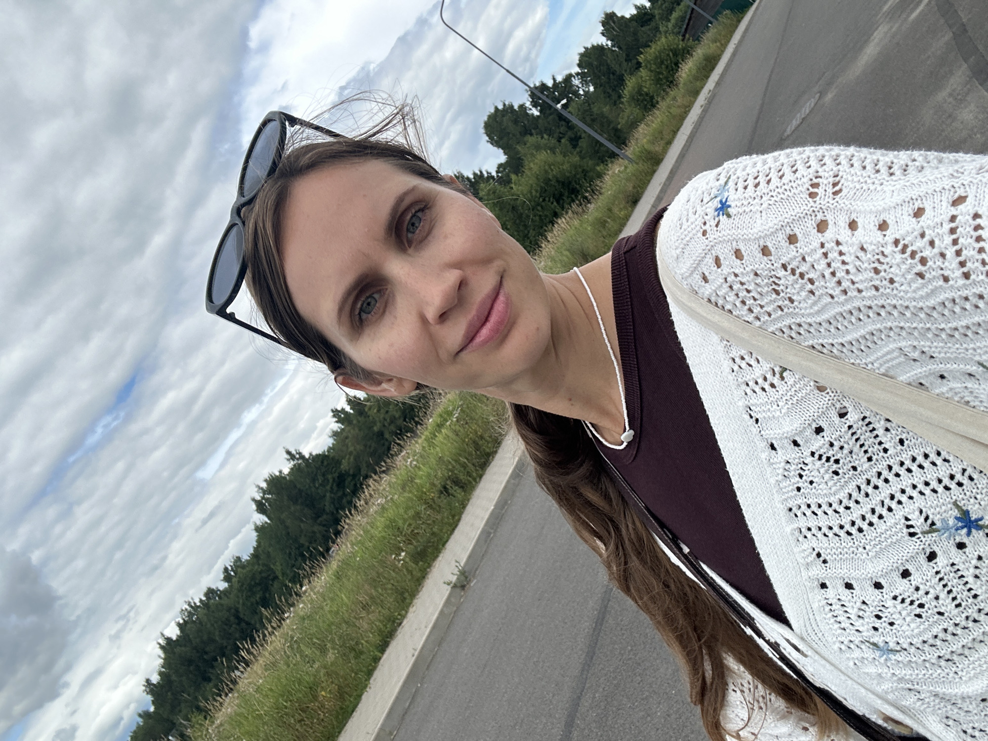 A medium shot, low-angle selfie captures a woman standing outdoors on a paved surface, likely a road or sidewalk. She has long, dark brown hair that falls over her left shoulder, and she is wearing a dark maroon top beneath a white open-knit cardigan. The cardigan has intricate patterns and small blue embroidered flowers on the right sleeve. She has a delicate white beaded necklace with a small pendant. Dark sunglasses are perched on top of her head.
The background consists of a grassy embankment with tall, dry grasses and scattered trees, suggesting a natural or park-like setting within a city. A gray paved road stretches out behind her, with a white curb separating it from the grassy area. In the distance, a lamppost is visible on the right side of the frame. The sky is overcast with dramatic clouds, hinting at variable weather conditions. The overall scene suggests a casual outdoor activity, possibly a walk or a moment of rest during a journey. The image does not contain any specific landmarks or architectural features that would definitively pinpoint the location within Vsevolozhsk, Russia, beyond a general sense of suburban or semi-rural environment.
