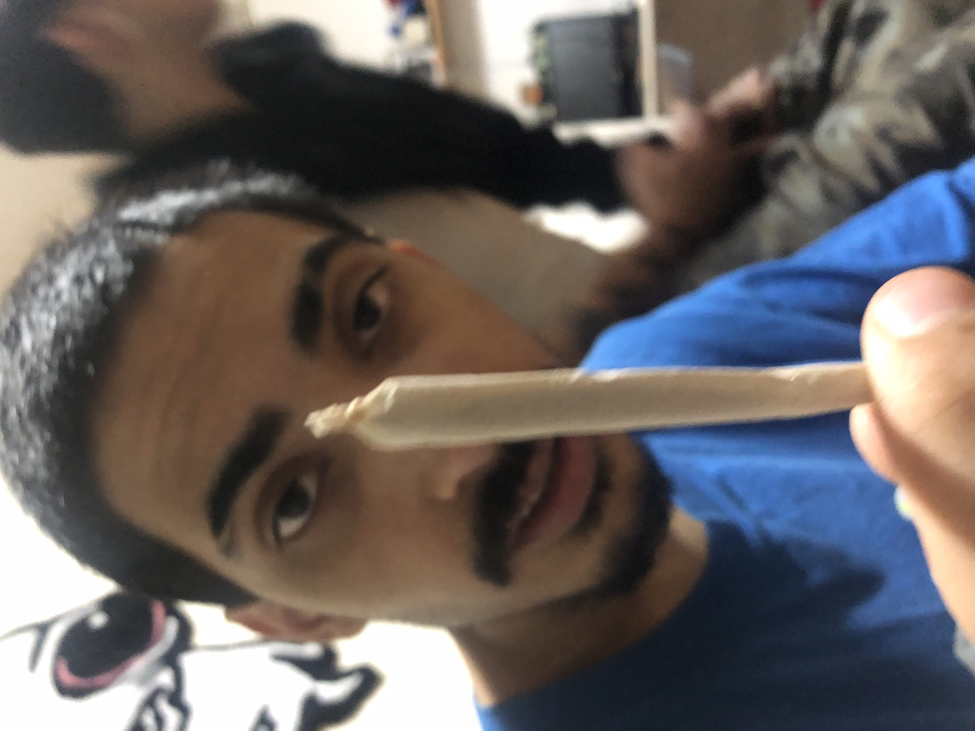 A close-up, angled shot shows a person holding a rolled cigarette in front of their face. The person has dark hair, dark eyebrows, and a dark mustache and goatee. They are wearing a blue t-shirt. Behind them, another person is partially visible, wearing a dark-colored shirt and camouflage pants. The background is blurry but includes a shelf or cabinet. The location is not discernible from the image.