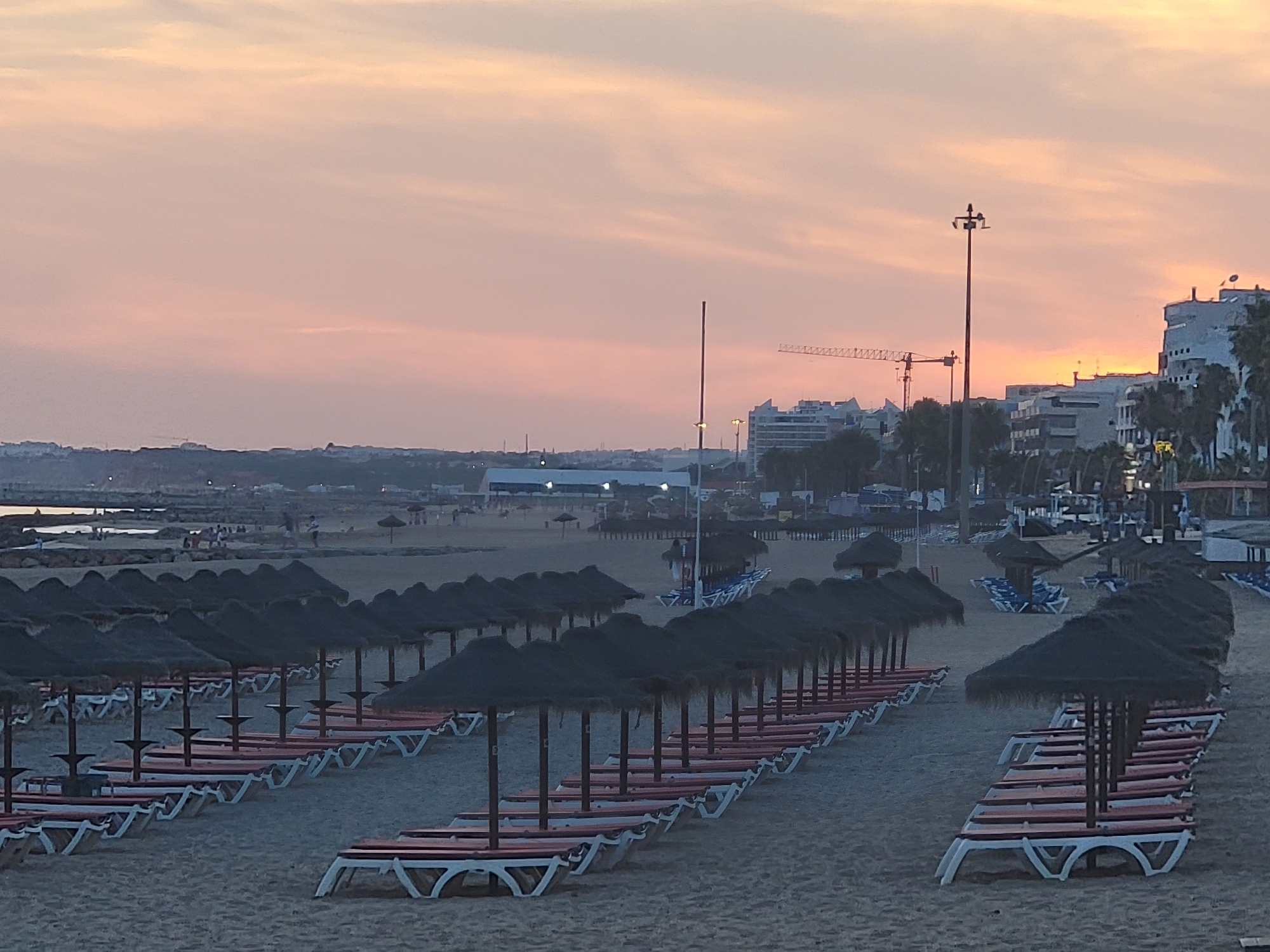 This is a photograph taken at dusk on a beach in Quarteira, Portugal. The sky is a soft pink and orange, indicating the setting sun. Rows of empty lounge chairs with orange cushions and thatched umbrellas are neatly arranged on the sandy beach. In the background, there are buildings, palm trees, and construction cranes, suggesting an urban environment adjacent to the coastline. Some people are visible in the distance, walking near the water's edge and sitting on the sand. The overall atmosphere is peaceful and serene.