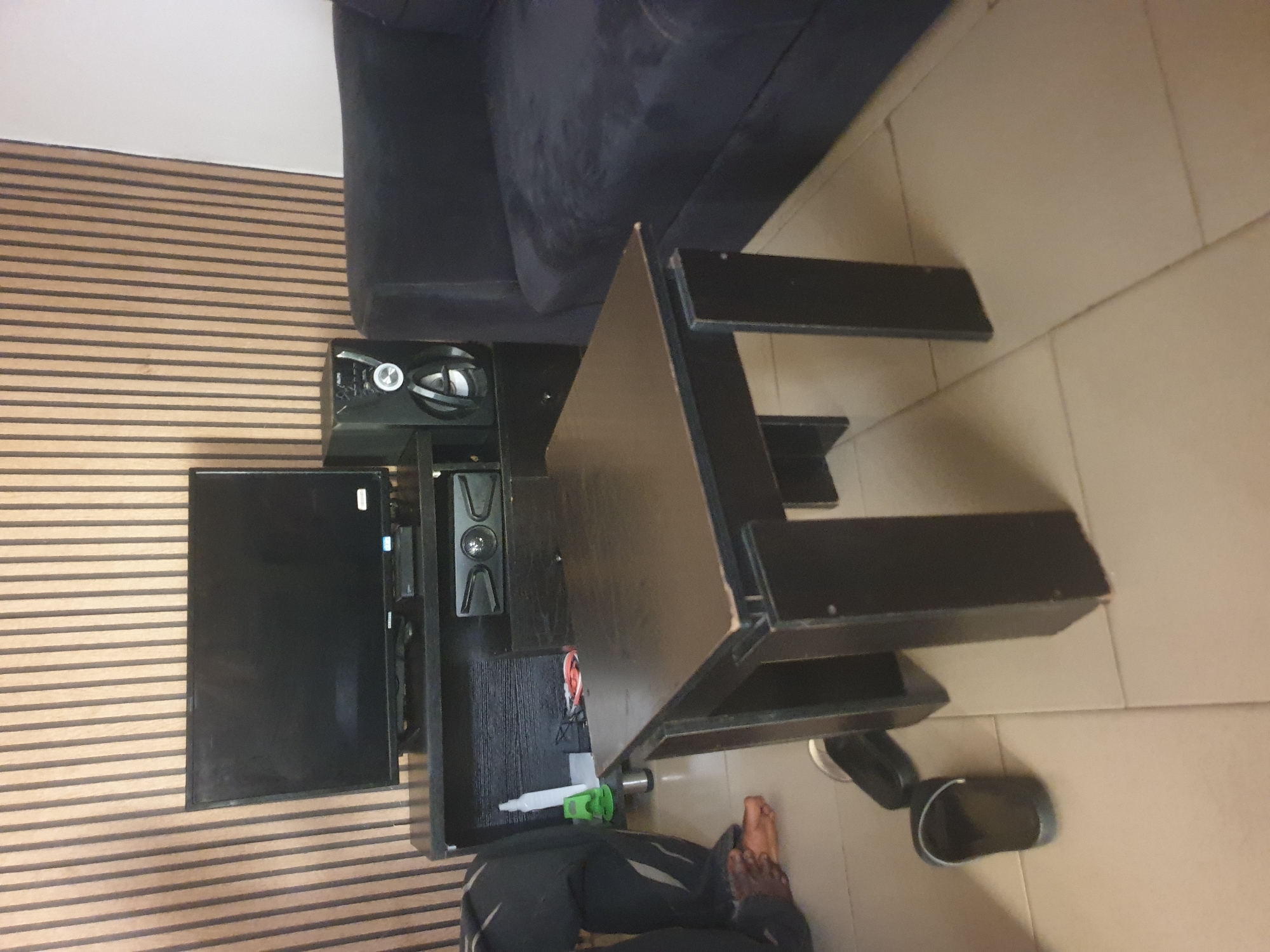 The image shows a living room scene with a dark wood coffee table in the foreground, positioned on a beige tiled floor. Behind the coffee table, a dark entertainment stand holds a flat-screen television and a speaker. To the right of the entertainment stand, a dark blue couch is visible. A person wearing dark pants is partially visible on the left side of the image, with their feet and legs showing, and they appear to be sitting or kneeling. A pair of black sandals is on the floor near the coffee table. The background features a wall with vertical wooden slats.