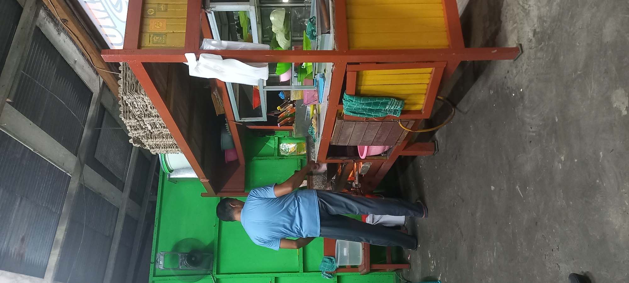 A man wearing a blue t-shirt and grey pants is standing in front of a food stall. The stall is made of wood with orange and yellow accents and has shelves stocked with various items. The man appears to be cooking or preparing food. The background is a green wall with a fan mounted on it. The floor is concrete. The scene suggests an outdoor or semi-outdoor food establishment in Loa Lepu, Indonesia.