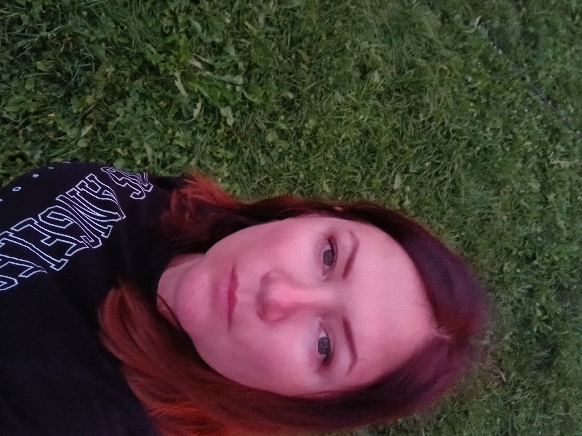 A woman with medium-length auburn hair is taking a selfie outdoors. She is wearing a black t-shirt with white lettering that appears to read "LOS ANGELES." The background consists of green grass. The image was taken at an unspecified time and location, and no connection to "Большая Уса, Russia" can be established from the visual information provided.