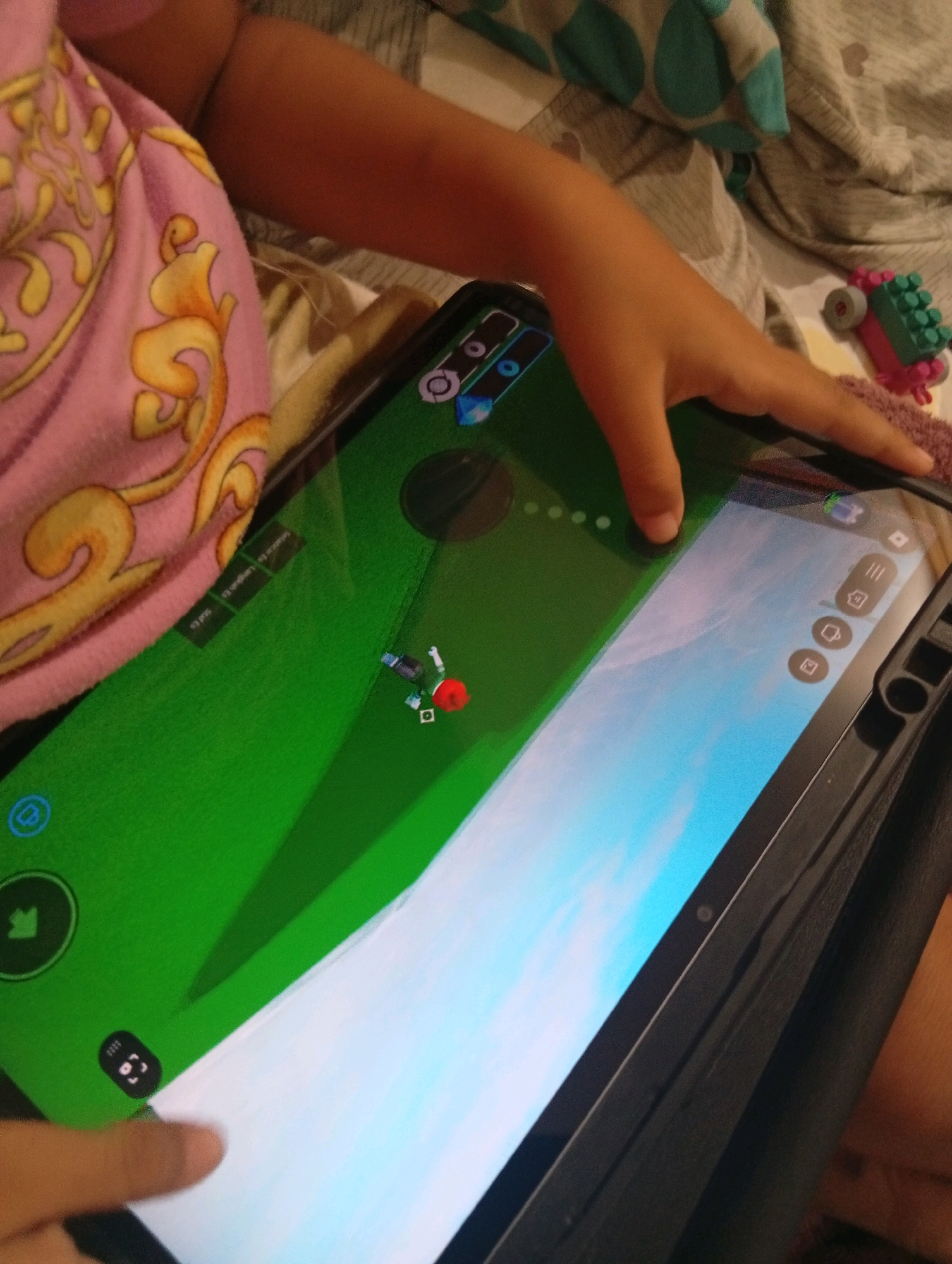A person's hand is visible in the foreground, interacting with a tablet screen. The tablet displays a video game with a green landscape and cartoon-like characters. In the background, a pink blanket with a gold pattern is draped over what appears to be a bed. There is also a portion of the person's arm and leg visible. A few toy building blocks are scattered in the background. The scene is likely indoors, possibly in a bedroom.
