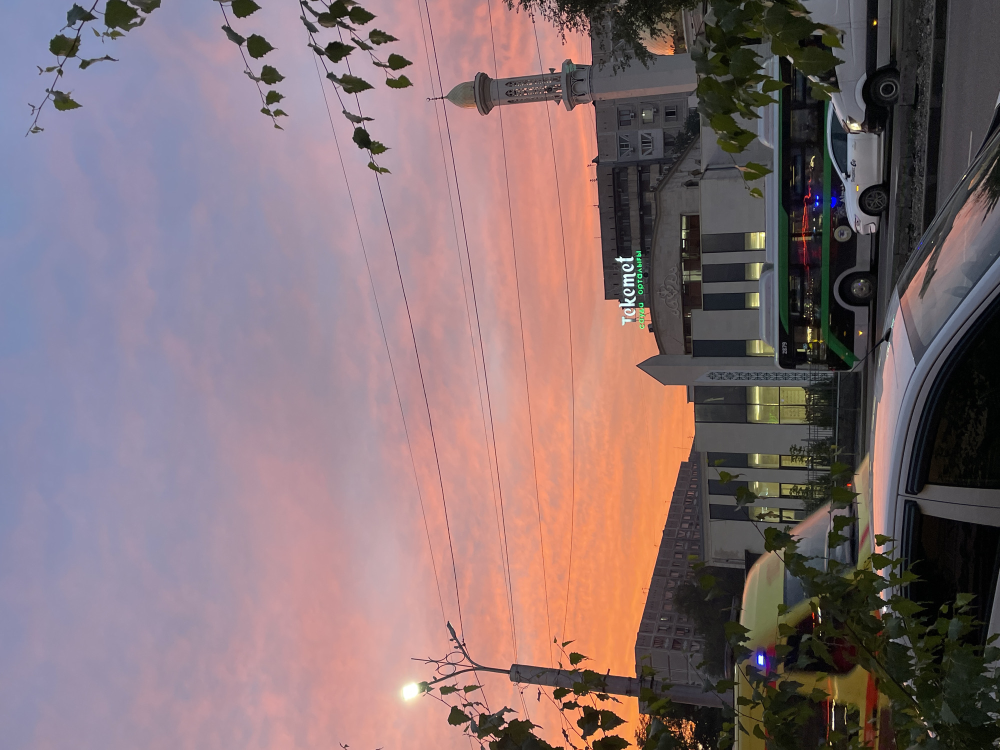 The image depicts a street scene in Almaty, Kazakhstan, during a sunset. The sky is ablaze with hues of pink and orange, with wispy clouds scattered across it. In the foreground, a street light illuminates the scene, with tree branches framing the composition on the left and right.
In the middle ground, several buildings are visible. A prominent building features the illuminated sign "Tekemet," which appears to be a business or shopping center. Adjacent to it, a minaret of a mosque stands tall against the vibrant sky. Traffic is evident on the street, with a bus, an ambulance, and several cars captured in motion. The bus is green and black with illuminated windows, and an ambulance with flashing blue lights is also visible. The reflections of the sunset can be seen on the surfaces of the vehicles.
The environment suggests an urban setting, with the presence of multi-story buildings, a street, and traffic indicating a populated area within Almaty. The time of day is clearly evening due to the sunset. There are no people clearly visible in the image, though the presence of vehicles implies human activity. The overall impression is a picturesque urban landscape enhanced by the dramatic colors of the sunset.