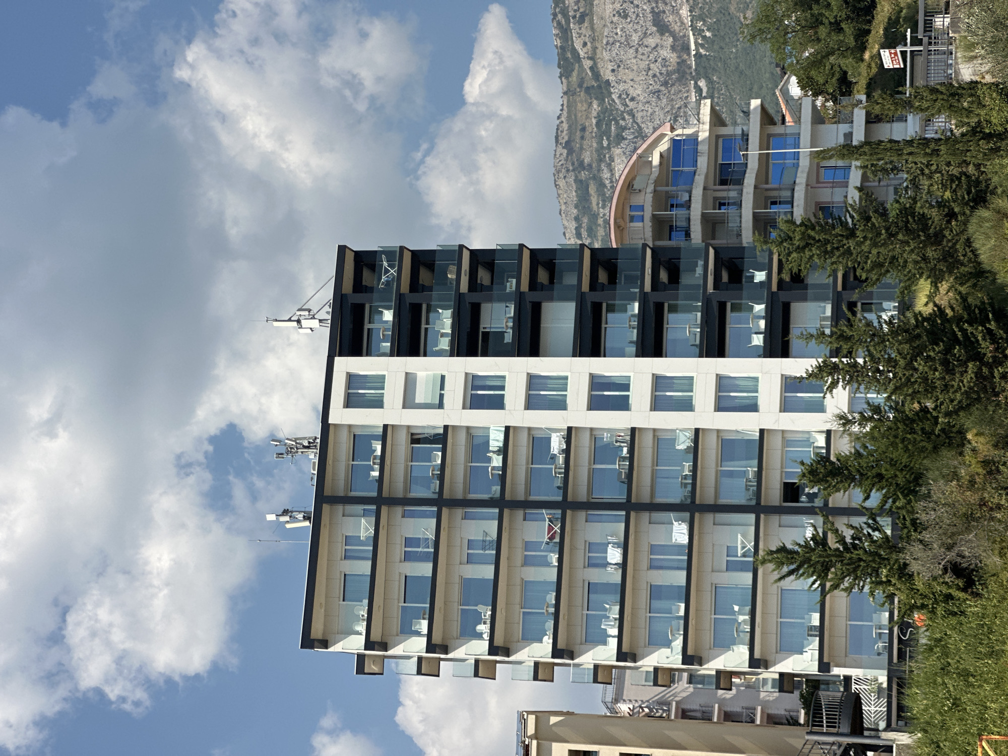 A medium-shot, eye-level, vertical, outdoor, daytime photograph shows a modern, multi-story hotel building on a hillside in Boreti, Montenegro. The hotel, with its white and dark grey facade, features numerous balconies with glass railings. Several balconies display white patio furniture, and one has laundry drying. The hotel has communication antennas on its roof. Adjacent to this hotel, on the left, is a portion of another building, a light-colored structure with balconies. Behind the hotels, a rugged, rocky mountain range rises under a blue sky filled with scattered white clouds. In the foreground, lush green trees and foliage obscure the lower portion of the buildings. A brown and white directional sign with red arrows is visible on the right side of the image, positioned near a street.