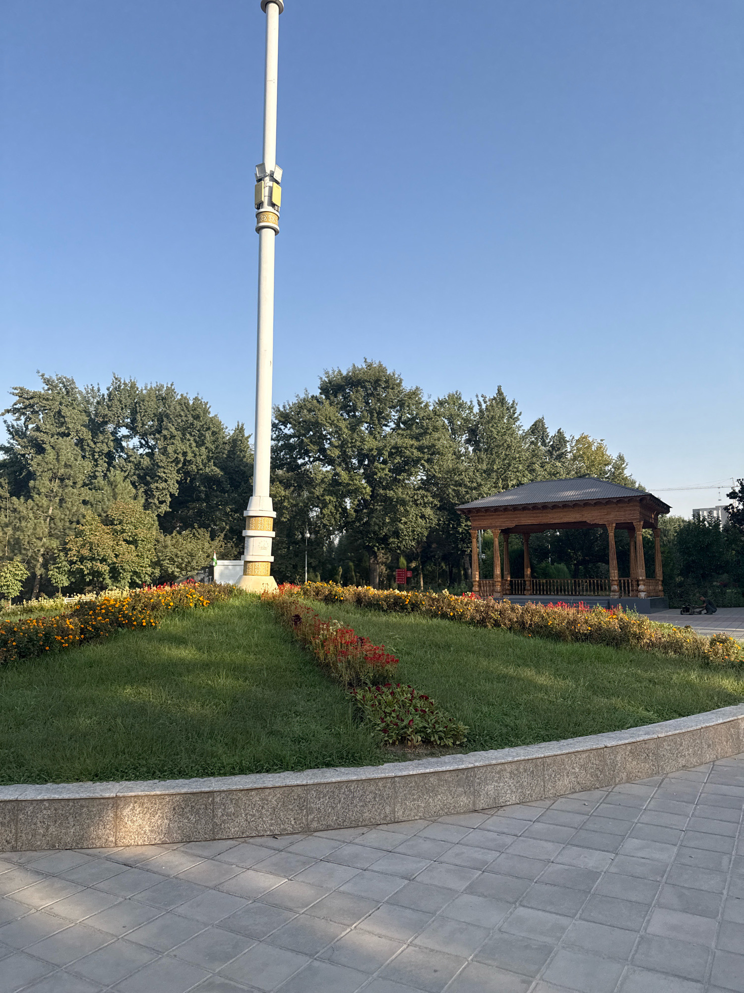 A tall, white lamppost with decorative bands stands in the center of a well-maintained park in Dushanbe, Tajikistan. The lamppost is surrounded by a circular, raised flower bed filled with a mix of orange, yellow, and red flowers. Behind the flower bed, a lush green lawn is visible, leading towards a cluster of mature trees. To the right of the lamppost and flower bed, a wooden gazebo with an ornate roof and carved details offers a shaded resting area. The ground in the foreground is paved with light gray square tiles. The sky is clear and blue, indicating fair weather. A person is partially visible sitting on the paved area to the right of the gazebo.
