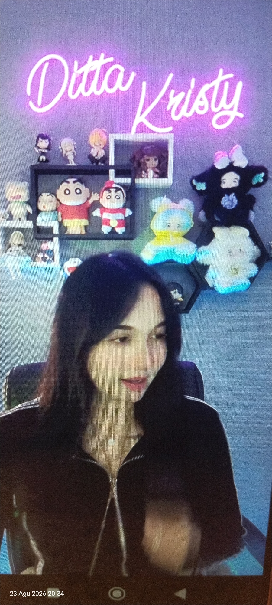 The image shows a woman with long dark hair and a black zip-up jacket, wearing a necklace. Behind her is a wall with a neon sign that reads "Ditta Kristy." There are shelves with various figurines and stuffed toys on the wall. The bottom left of the image shows a timestamp: "23 Agu 2026 20.34". The background appears to be an indoor setting, likely a room. The quality of the image is somewhat blurry, suggesting it was taken with a phone camera in low light conditions. There is no identifiable information about the specific location within East Jakarta, Indonesia.