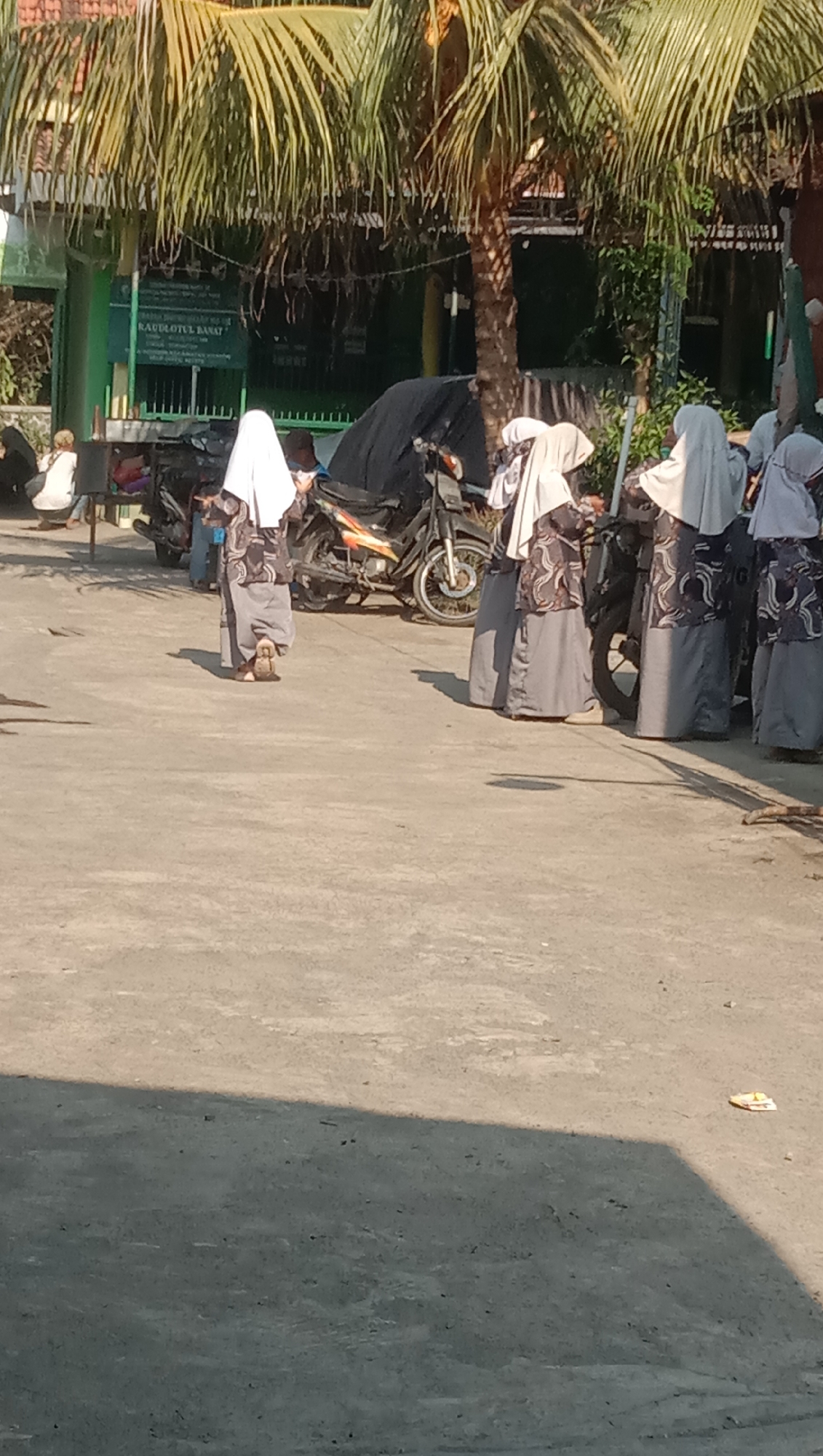 The image depicts a sunny outdoor scene, likely a schoolyard or courtyard, in Sidogiri, Indonesia. In the foreground, a group of five young girls, dressed in traditional Islamic attire with hijabs and long patterned tunics over grey skirts, are walking or standing. The girl closest to the camera is facing away, showing her back and the sole of her left shoe. Behind her, four other girls are also in similar attire, some are looking towards the viewer, others away. Their clothing features a repeating pattern of grey and white abstract shapes.
To the left of the group, a person in white clothing is sitting on a low structure, possibly a bench, near a green building. Behind the girls, several motorcycles are parked. One motorcycle, in the center of the frame, is black with orange and yellow accents. In the background, a green building with a sign that reads "RAUDLOTUL BANAT" is visible, suggesting it might be an educational institution. A palm tree with large fronds towers over the scene, casting a shadow across the paved ground. The ground itself is a light grey, uneven surface, with some scattered debris like a small piece of yellow and white plastic. The overall impression is a candid snapshot of daily life in a school environment, under bright sunlight.