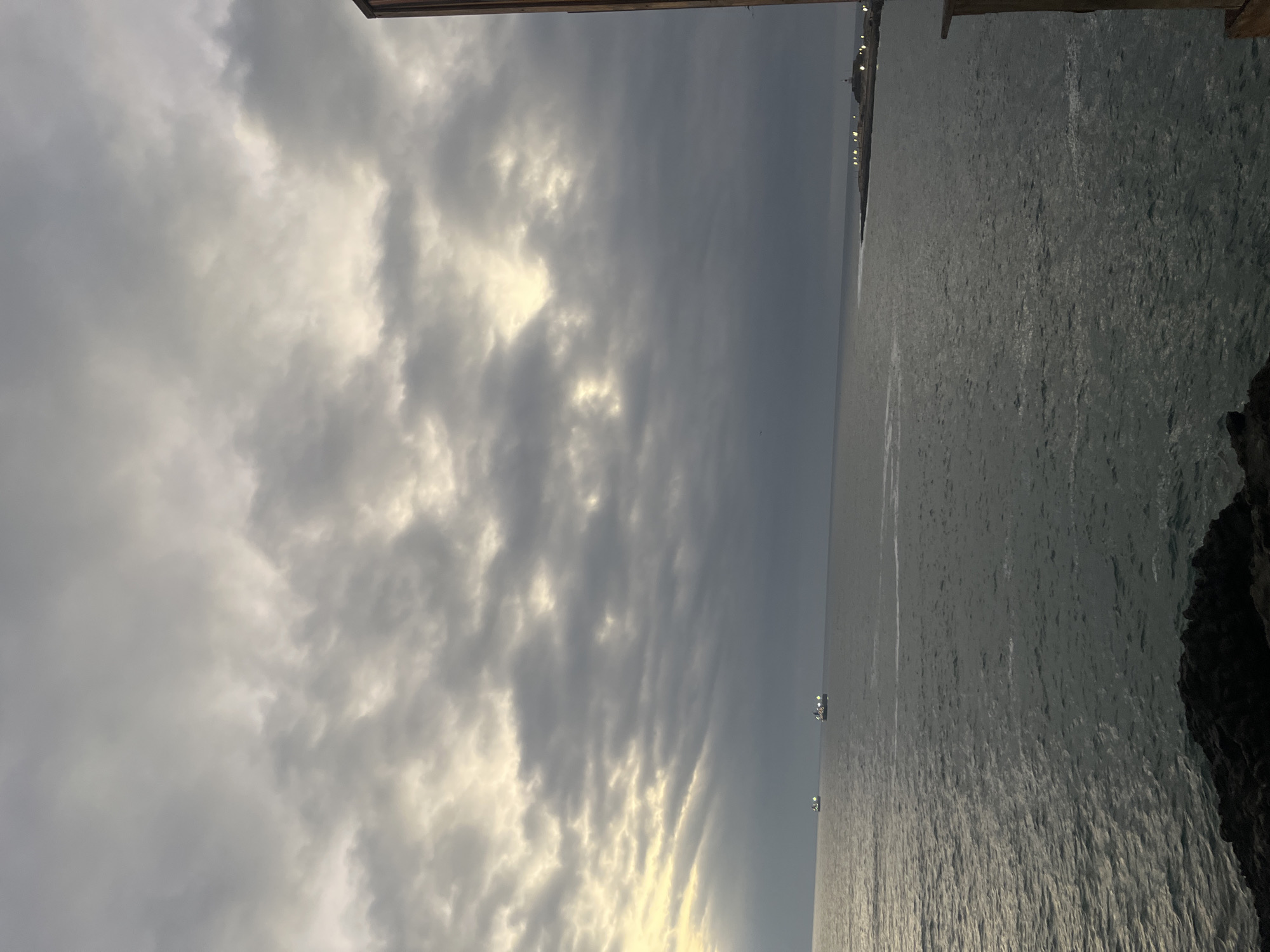 The image depicts a view of the Pacific Ocean in Arica, Chile, under a cloudy sky. The sky is predominantly grey with lighter areas suggesting sunlight breaking through the clouds. The water is dark with subtle reflections, hinting at a choppy surface. At the edge of the water is the black, rocky edge of the coastline. In the background a series of lights are faintly visible.