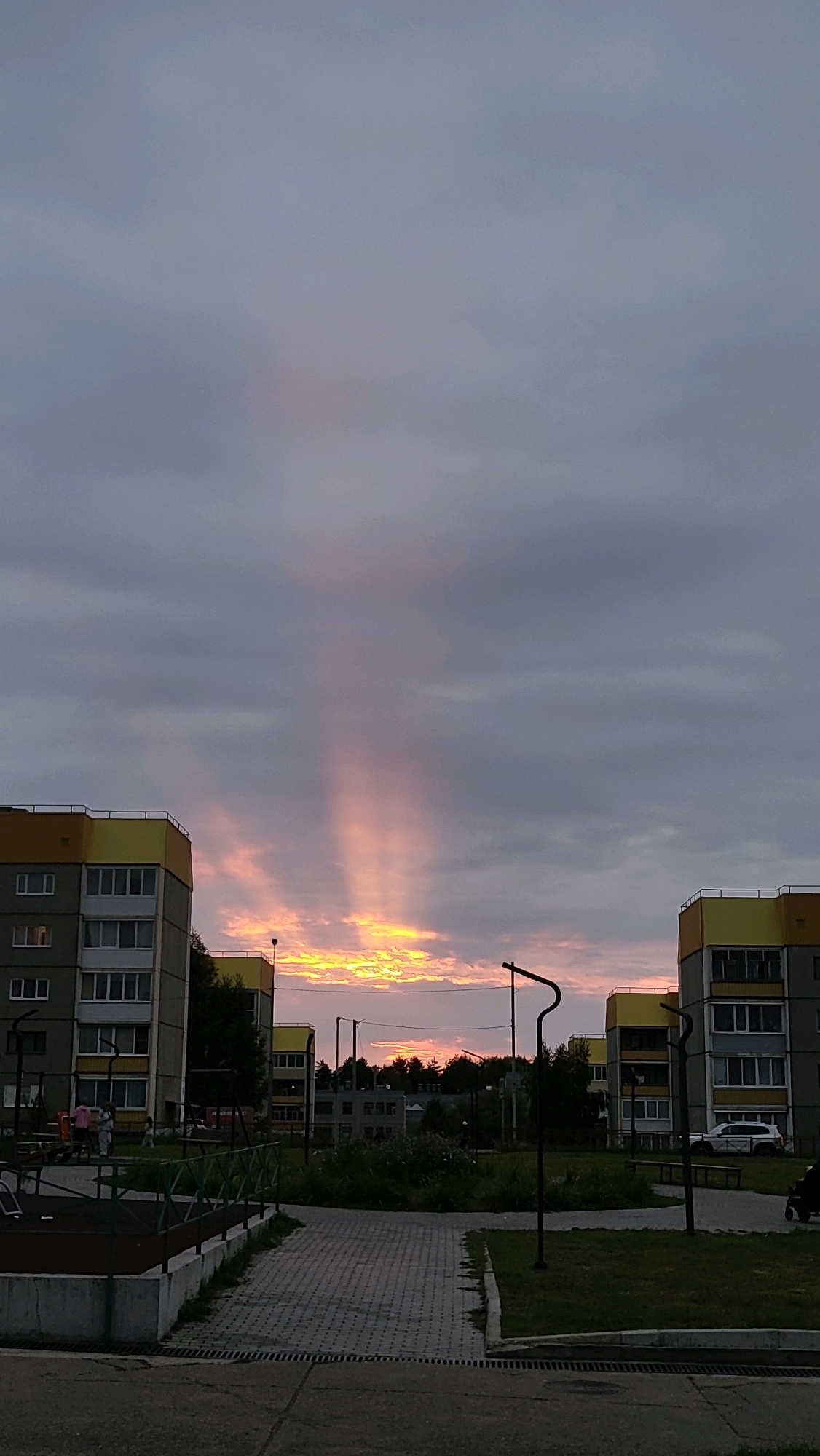 This is a vertical, eye-level shot taken from a paved area looking towards a residential courtyard in the city of Markovskiy, Russia, during twilight. The sky is filled with a gradient of grey clouds, with a distinct shaft of pink and yellow light beaming down from the horizon, indicating a sunset or sunrise through a break in the clouds. In the foreground, a paved pathway leads into the courtyard. To the left and right, there are multi-story apartment buildings with yellow and grey facades. Several trees are visible scattered throughout the courtyard, as well as streetlights and benches. There are a few people in the distance, near a playground area on the left, and a white SUV parked near the building on the right.