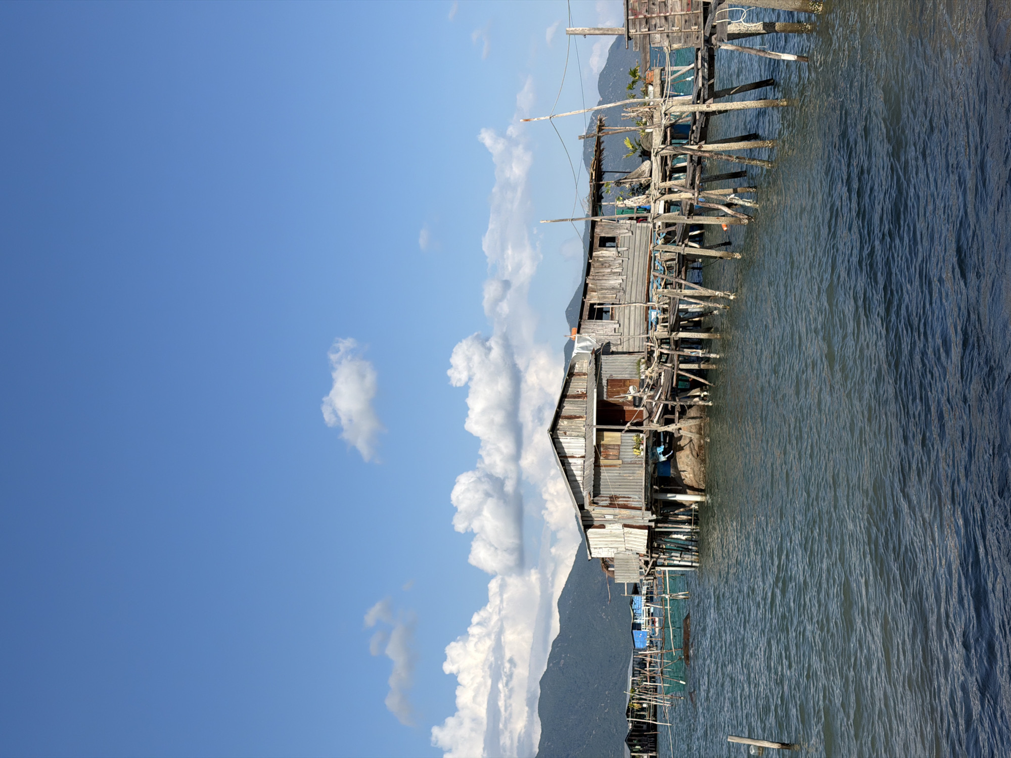 Captured in Bac Nha Trang Ward, Vietnam, the image portrays a wooden structure built over water, supported by stilts. The architecture appears aged, with weathered materials. The water's surface is visible, indicative of a coastal or lakeside setting. A mountain is situated in the background. The sky is blue with clouds, suggesting daylight. The photo's composition includes elements of nature and man-made structures, indicative of human habitation in a waterfront environment.