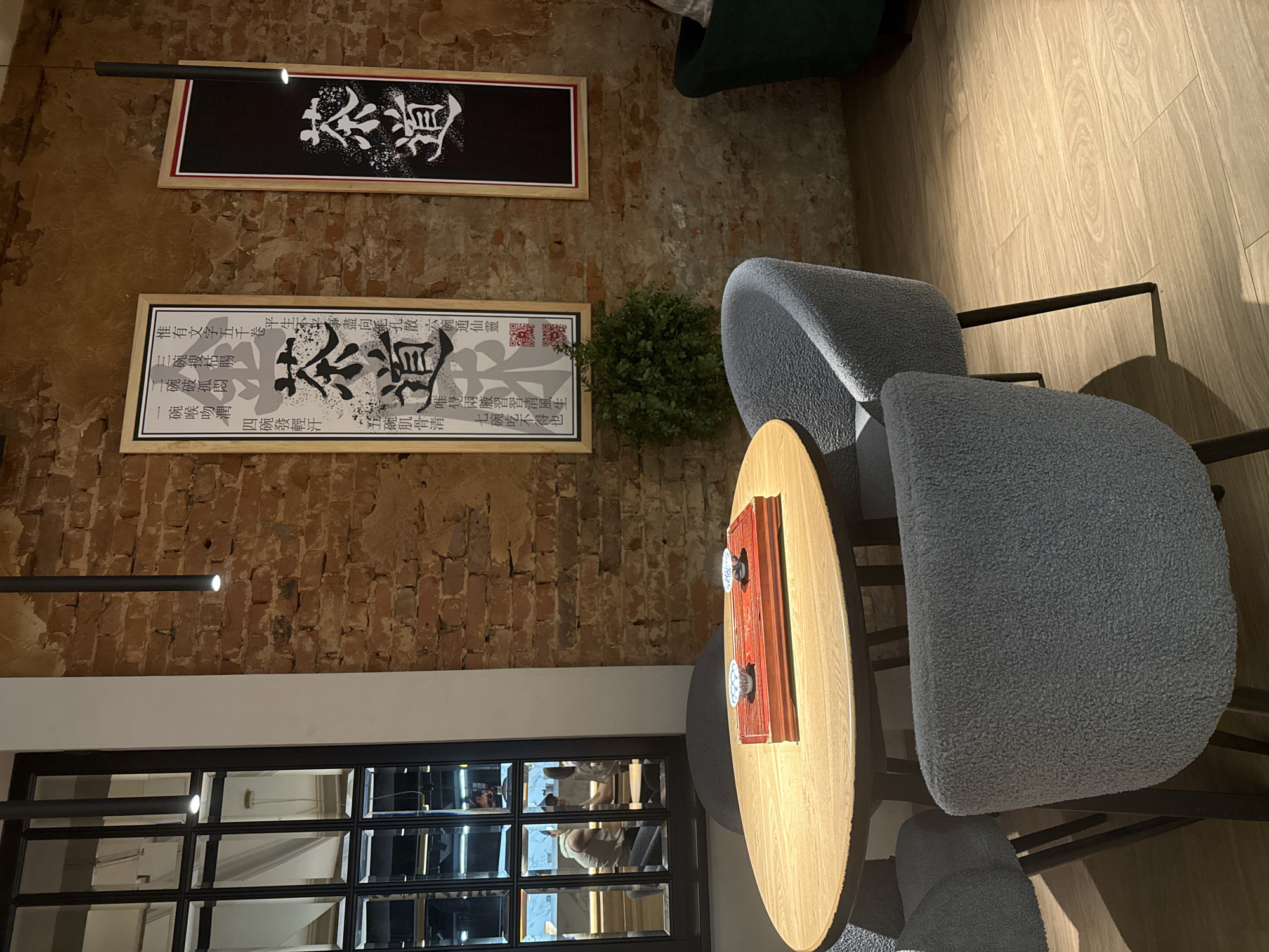 The image depicts an interior view of what appears to be a cafe or tea house. In the foreground, a round wooden table with dark legs occupies the central space, surrounded by four grey, textured, upholstered chairs, also with dark legs. Placed on the table is a reddish-brown rectangular wooden tray holding two small, white ceramic cups with blue patterns.
To the left, a large, black-framed window with multiple panes reflects another section of the interior. Within this reflection, at least two individuals are visible; one male in a light-colored sleeveless top is seated, with another person partially obscured beside him, suggesting patrons at a counter or another table. A white vertical wall section separates the window from the main background wall.
The background features an exposed brick wall, displaying varied shades of red, orange, and brown, with visible mortar. Two framed artworks hang on this wall. The left artwork is a white poster with black and grey Chinese calligraphy, prominently featuring the characters "茶道" (Chádào - The Way of Tea), along with smaller red text and a QR code, all enclosed in a light wooden frame. The right artwork is a black poster with white Chinese calligraphy, also displaying "茶道," set within a light wooden frame with a reddish inner border. Below the left poster, a small, densely foliated green potted plant sits on the floor. A partial view of a dark green upholstered armchair or sofa is visible in the far right corner.
Several black, cylindrical pendant lights hang from the ceiling, one directly above the foreground table and another further back, providing focused illumination. The floor is covered with light-colored wooden planks exhibiting distinct grain patterns. The overall scene suggests a cultivated, perhaps East Asian-themed, casual dining or relaxation environment.