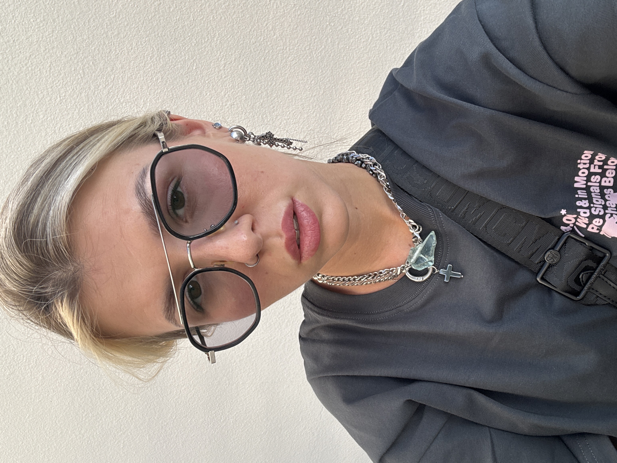 A close-up, eye-level, medium shot captures a young woman from the shoulders up, against a textured, off-white wall. She is wearing large, round, black-rimmed glasses with a silver double bridge, and her blonde hair is pulled back. She has light-colored eyes, a small nose ring, and her lips are slightly parted, painted with pink lipstick. She wears a silver necklace with a large, irregular pale blue pendant and a smaller silver cross pendant, layered over a dark gray t-shirt. A black strap with a silver buckle is visible across her chest. She has multiple piercings in her ears, including a dangling silver earring with multiple charms. The lighting is even, and the background is neutral. The scene appears to be outdoors or in a well-lit indoor space.
