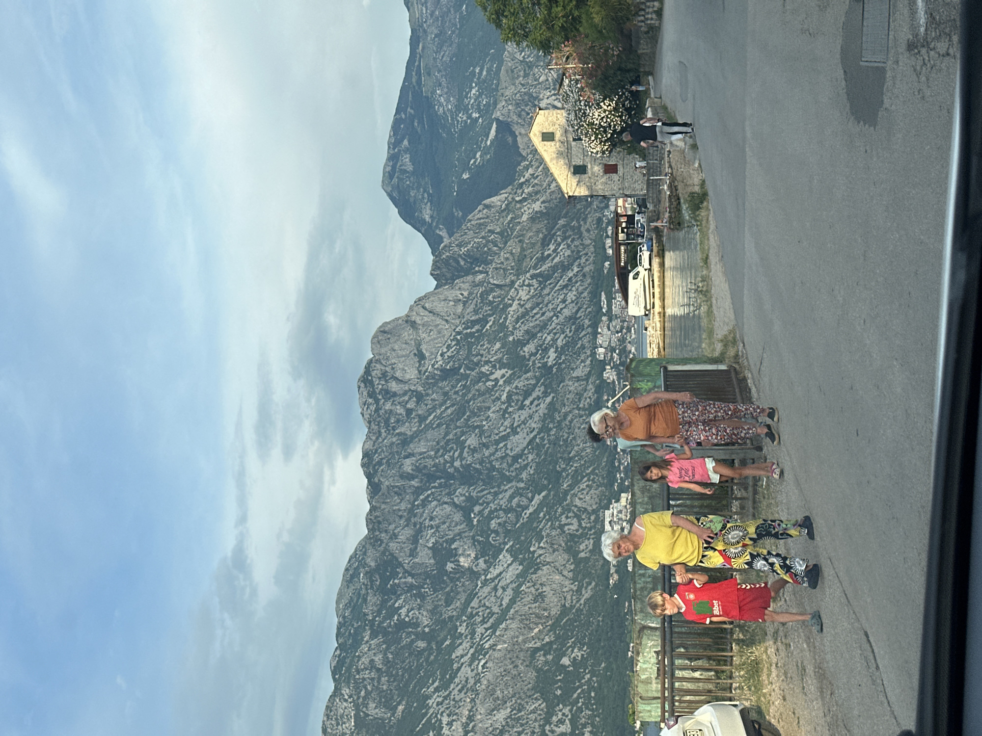The image shows a group of people standing on a paved road with a steep, rocky mountain in the background. The mountain is covered in sparse vegetation and appears to be a prominent feature of the landscape. In the mid-ground, there is a building with a light-colored facade and a red-tiled roof, surrounded by trees and foliage. Several cars are parked along the road. The sky is overcast with a mix of light and dark clouds, suggesting it might be late afternoon or early evening.
The people are positioned along the edge of the road, some appearing to be in conversation or observing the scenery. There are at least four individuals visible, including what appears to be an older woman wearing an orange top and patterned pants, a young girl in a pink t-shirt and white shorts, another older woman in a yellow top and vibrantly patterned pants, and a young boy in a red t-shirt and red shorts. The overall mood seems relaxed and casual.
The scene is set in Prčanj, Montenegro, a coastal town known for its Baroque architecture and its location against a backdrop of dramatic mountains. The presence of the road, buildings, and vehicles suggests a developed area. There is some visible text on the red t-shirt of the boy, which appears to be a logo or word.