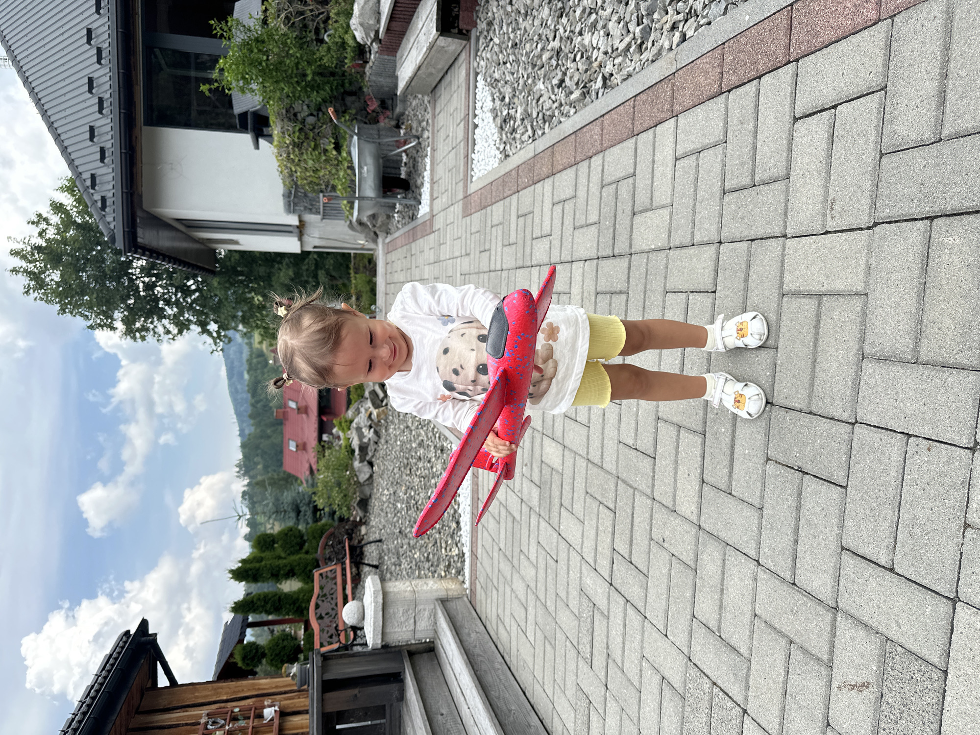 A young girl stands on a paved walkway holding a red toy airplane. She is wearing a white long-sleeved shirt with a picture of a dog on it and yellow shorts, along with white sandals. The background shows a house with a dark roof, trees, and a hillside. The scene appears to be in a suburban or rural setting, likely in the city of Wisła, Poland, given the architectural style and mountainous backdrop. The sky is partly cloudy.