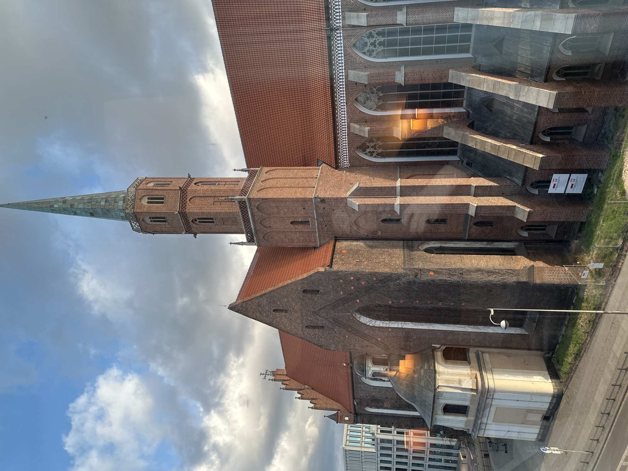 The image captures a side view of the Collegiate Church of the Holy Cross and St. Bartholomew, located in Wrocław, Poland. The architectural style is predominantly Gothic, characterized by the red brick construction, pointed arches, and high-pitched roofs. A slender tower with a green-tiled spire rises prominently. The exterior features buttresses and tall windows. In the foreground, a modern building with glass windows is partially visible. The sky is filled with clouds and patches of blue. A street and sidewalks are visible in the lower right corner. The image appears to have been taken through a window.