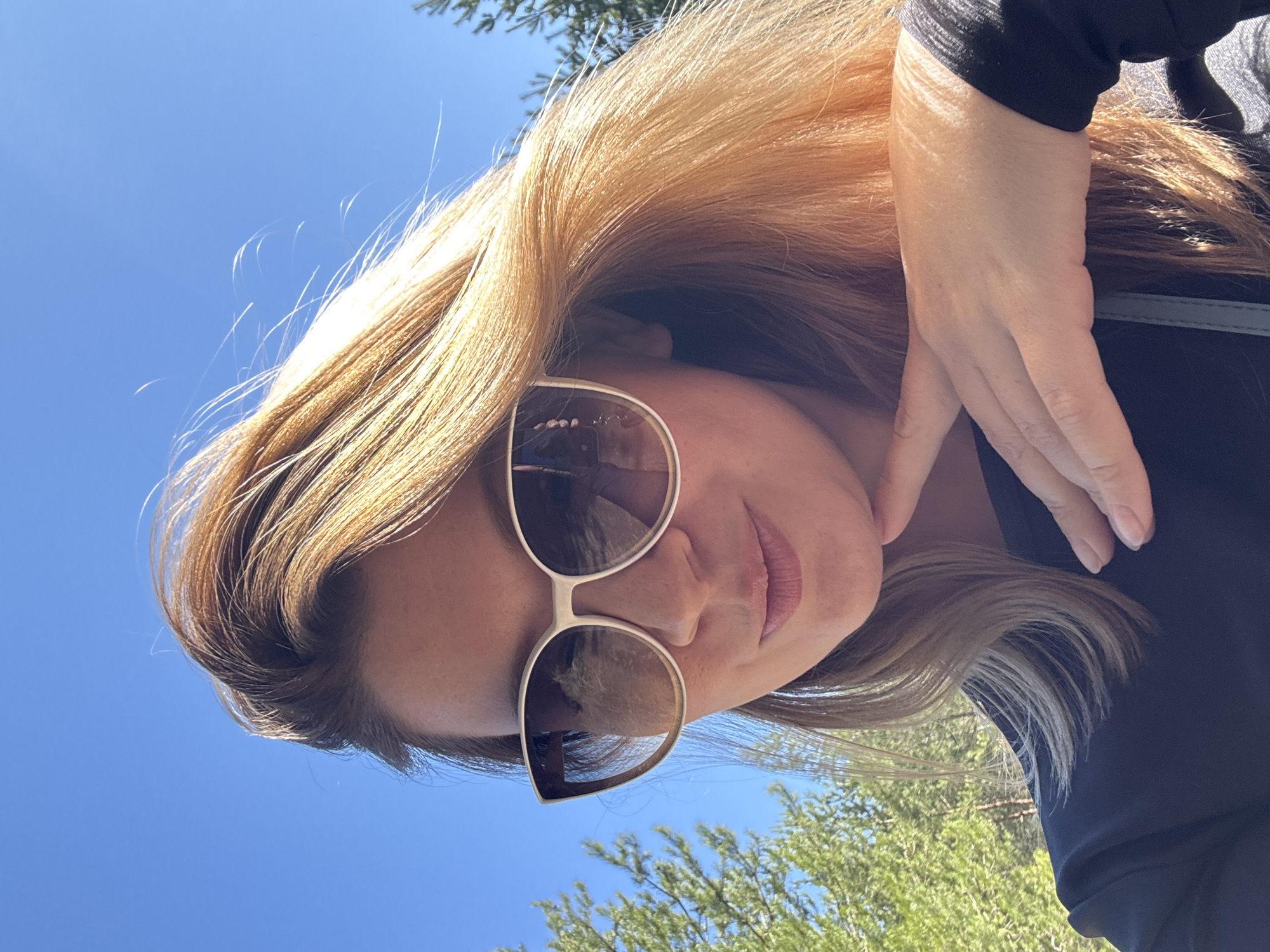 A woman wearing sunglasses and a black shirt is the primary subject of the image. Her hand is placed near her face. The background includes a blue sky and green foliage. The location is not identifiable as the city of Chaikovsky, Russia.