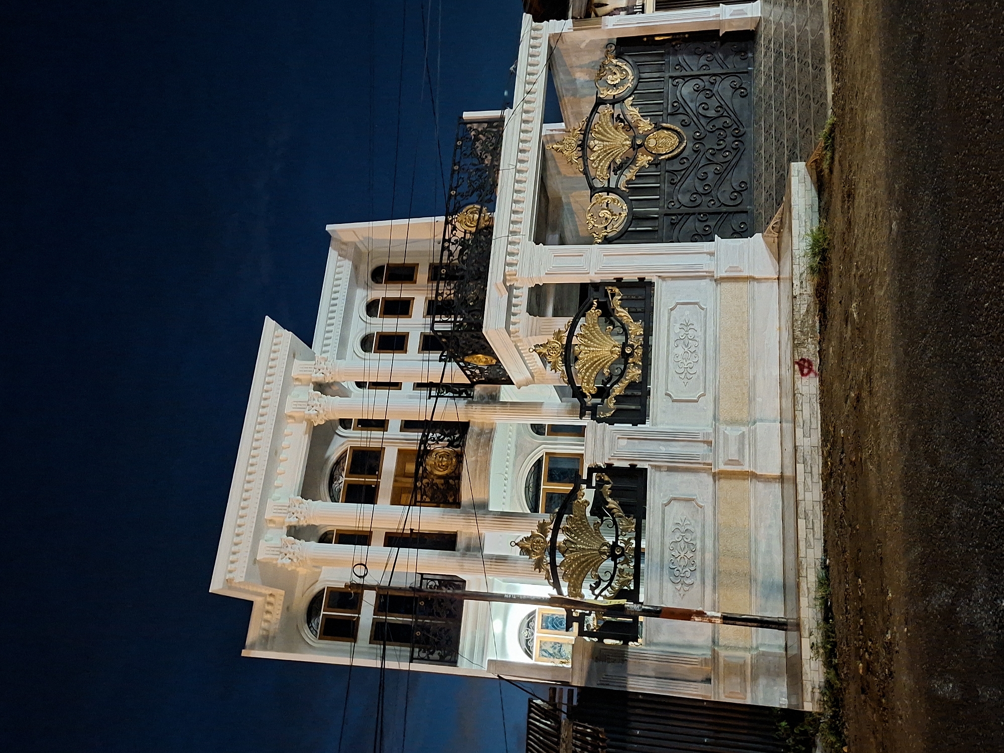 The image shows a multi-story building in Lhokseumawe, Indonesia, photographed at night. The building features white facades, ornate gold and black ironwork, and a classical architectural style with columns. The sky is a deep blue. The building is adjacent to a road. Vegetation grows at the edge of the road. Utility wires are visible.