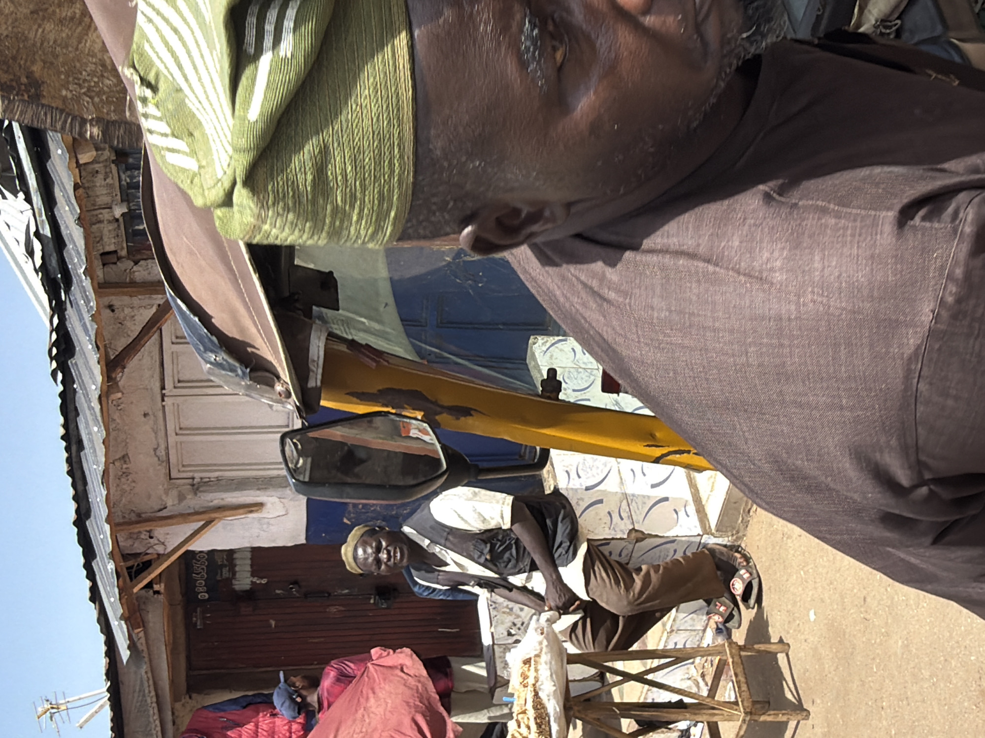 The image captures an outdoor street scene in Gura Top, Nigeria, under bright, clear daylight. The perspective is from a close, slightly elevated viewpoint.
In the immediate foreground, a person with dark skin is partially visible on the right side of the frame, seen in profile from the chest up. They are wearing a light green, ribbed head covering or scarf with white stripes, and a dark brown shirt or garment. Their right ear and a portion of their face are visible.
In the midground, slightly to the left, a man is seated on a low, rustic wooden stool. He has dark skin and is wearing a light-colored cap, a light short-sleeved shirt over a dark vest or jacket, and dark brown trousers paired with sandals. He looks towards the camera with a neutral expression, appearing to be engaged with a white bag or sack that rests on the stool before him, suggesting an activity like sorting or selling goods.
Behind the seated man, parts of a yellow vehicle, possibly a tricycle taxi or tuk-tuk, are visible, including its rearview mirror. The background features local architecture: a building with a corrugated metal roof, weathered concrete walls, and wooden support beams. A wall behind the seated man displays white and blue patterned tiles or paint, with a dark brown corrugated metal door or shutter below. A partially visible phone number, "080659080", is present on a surface near the yellow vehicle. In the lower left, a red fabric or blanket is visible, along with a glimpse of another person wearing a blue cap. An antenna stands out against the clear blue sky in the far background on the left. The scene depicts a candid moment of daily life and street commerce.