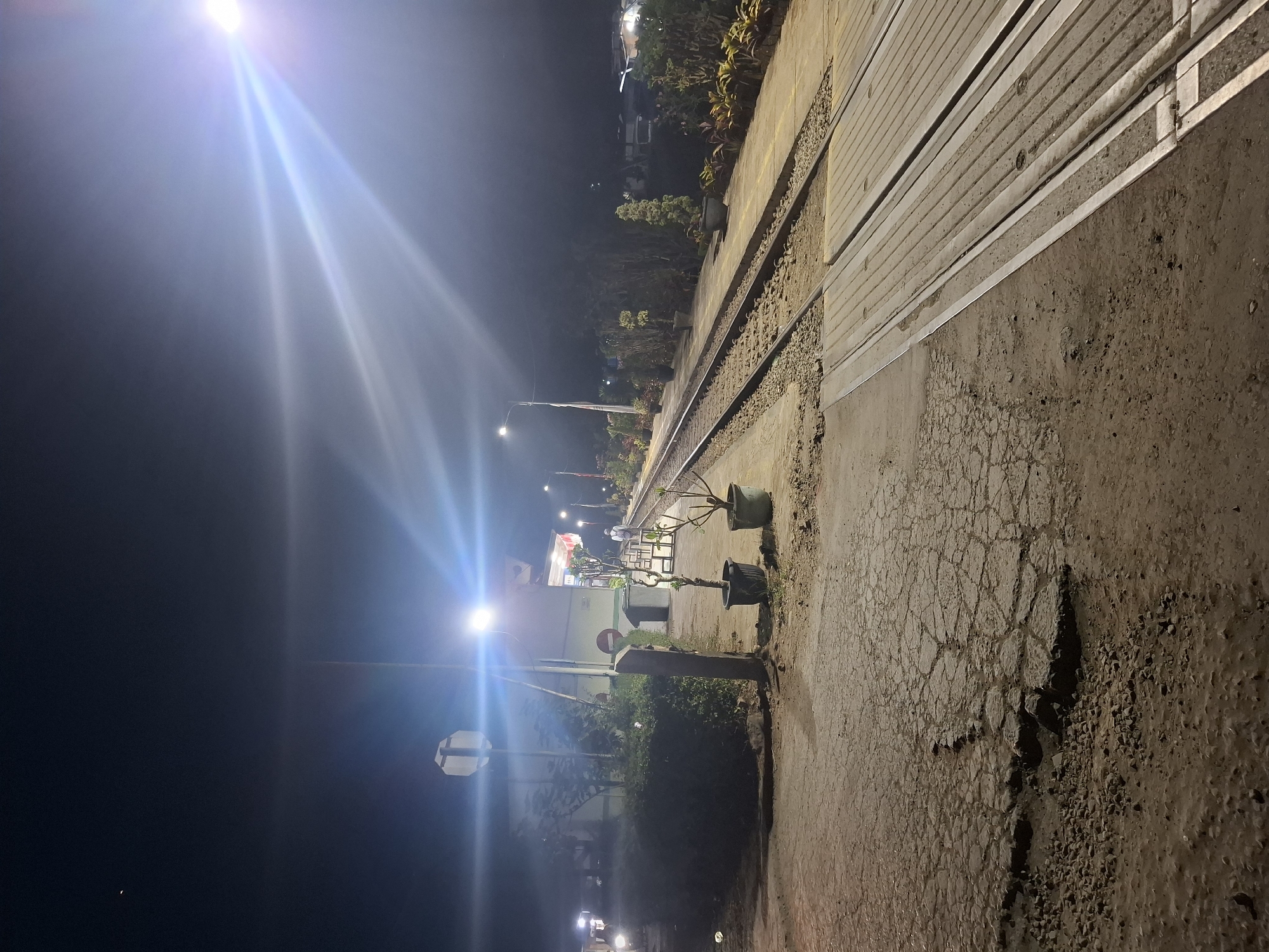 The image captures a nighttime scene at a train station in Ciheulang Tonggoh, Indonesia. The foreground shows a cracked, uneven dirt and gravel path leading towards a set of train tracks. The tracks run diagonally across the frame, illuminated by bright overhead lights that create strong lens flares. To the right of the tracks, a paved platform is visible, lined with potted plants. In the mid-ground, a small building, likely a station house, is present, with more lights and some vegetation. A person in white clothing is walking on the platform. To the left, a dense hedge lines the edge of the platform, and a motorcycle is partially visible in the distance. The overall atmosphere is dimly lit and somewhat desolate, with artificial lighting dominating the scene.