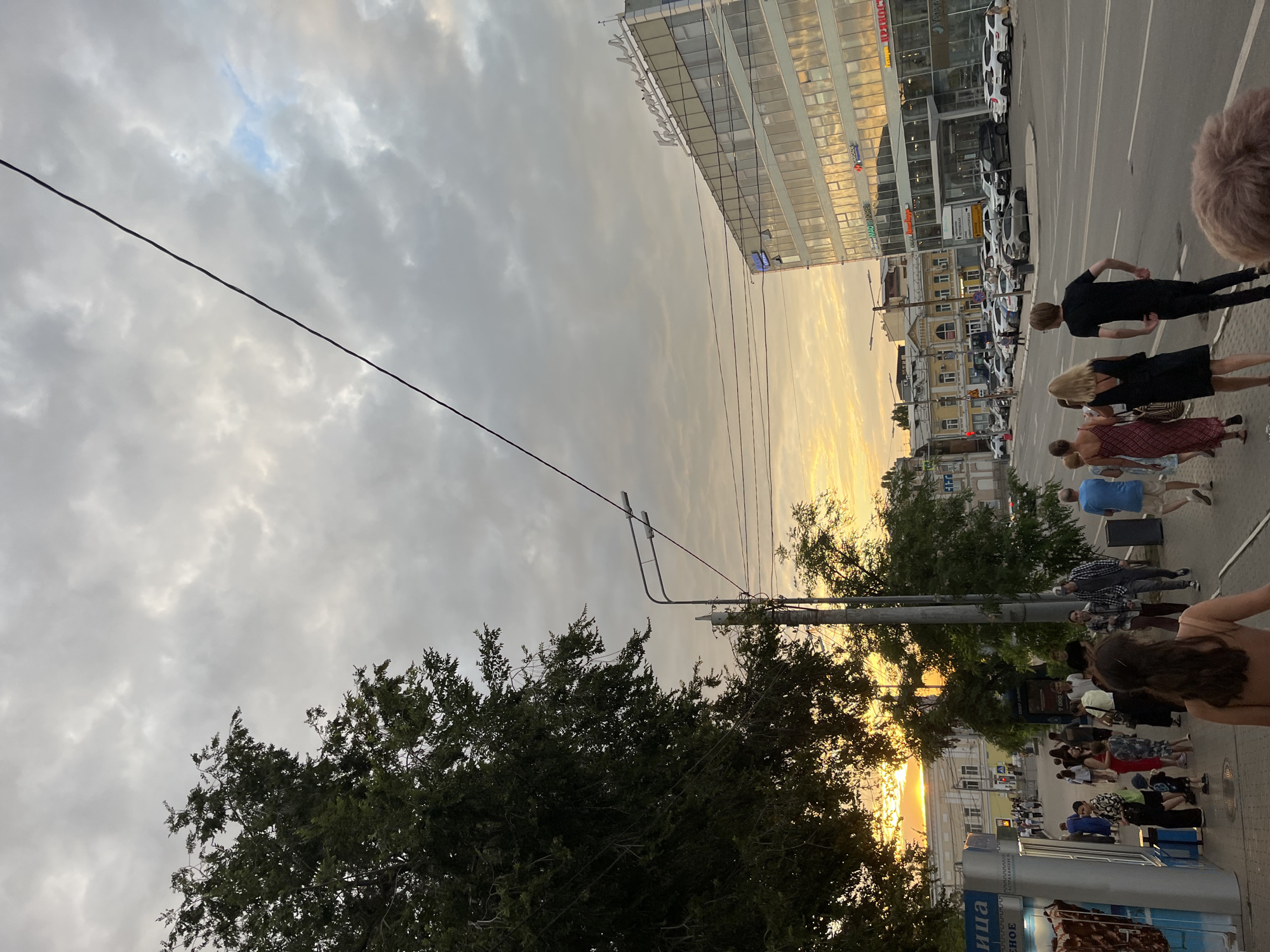 The image captures a street scene in Astrakhan, Russia, likely during the late afternoon or early evening, as indicated by the warm, golden hues of the sunset filtering through the clouds. On the right side of the frame, a modern, multi-story glass-fronted building dominates the skyline, reflecting the sky and surrounding cityscape. The building has signage with Cyrillic script, one of which appears to read "Кристалл" (Kristall). In front of this building, a parking lot is filled with various cars.
The left side of the image features a sidewalk lined with trees, casting shadows and partially obscuring a view of older, yellow buildings in the background. A street light with multiple lamps stands prominently near the center of the frame. A kiosk or small storefront with a blue sign displaying text in Russian is also visible on the left.
A diverse group of people are walking on the sidewalk, some singly and others in small groups. They are dressed in casual summer attire. One woman in the foreground is partially visible from behind, facing away from the camera. A group of four individuals, three women and one man, are walking together further down the sidewalk. Other pedestrians are scattered along the street. The sky is filled with dramatic, overcast clouds, with patches of brighter sky suggesting the setting sun. Electrical wires are strung across the upper portion of the image. The overall impression is one of a bustling urban environment bathed in the soft light of dusk.