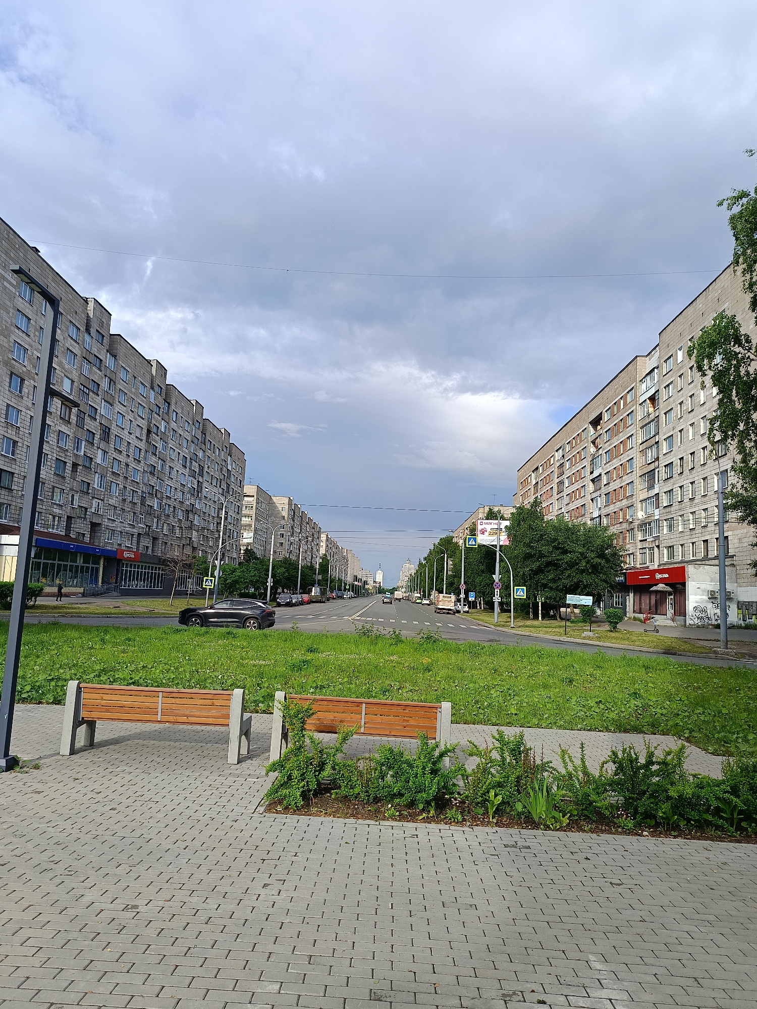 The image depicts a street scene in a city, with tall, grey apartment buildings lining both sides of a wide road. The buildings are made of concrete blocks and have many windows. A green grassy median separates the lanes of traffic, and trees are planted along the sides of the road. In the foreground, there is a paved walkway with two wooden benches. A few cars are visible on the road, and there are traffic signs and streetlights. The sky is overcast with grey clouds. The environment appears to be a residential area with commercial establishments on the ground floors of the buildings. The time of day is likely daytime, given the lighting conditions.
