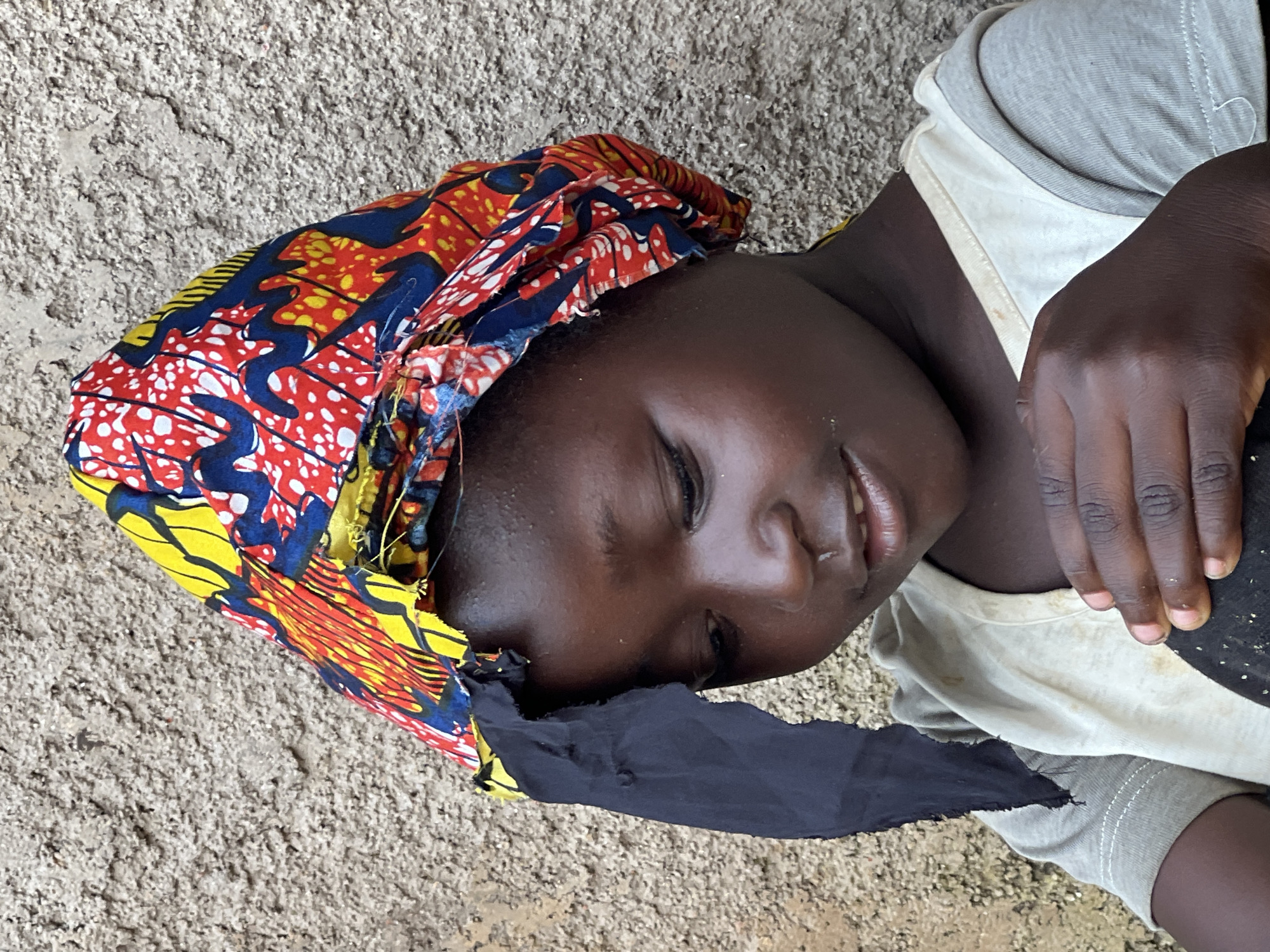 This is a close-up, slightly angled outdoor photograph of a young Black person lying down. They are wearing a vibrant, patterned headscarf with colors of red, yellow, blue, and white, which partially covers their head and forehead. A piece of dark fabric is draped over their face, obscuring the lower half. The person is wearing a light-colored t-shirt with grey accents. Their visible hand is resting on their chest, with fingers slightly curled. The background appears to be a rough, textured surface, possibly the ground or a wall, in a light brown or grey color. The lighting suggests it is daytime, and there are no strong shadows, indicating possibly overcast conditions or diffused sunlight. The scene has a candid feel, and the person's eyes are closed, with a faint smile suggested by the visible part of their mouth, conveying a sense of peace or rest. The location context of Jos, Nigeria, places the image in a specific geographical and cultural setting.
