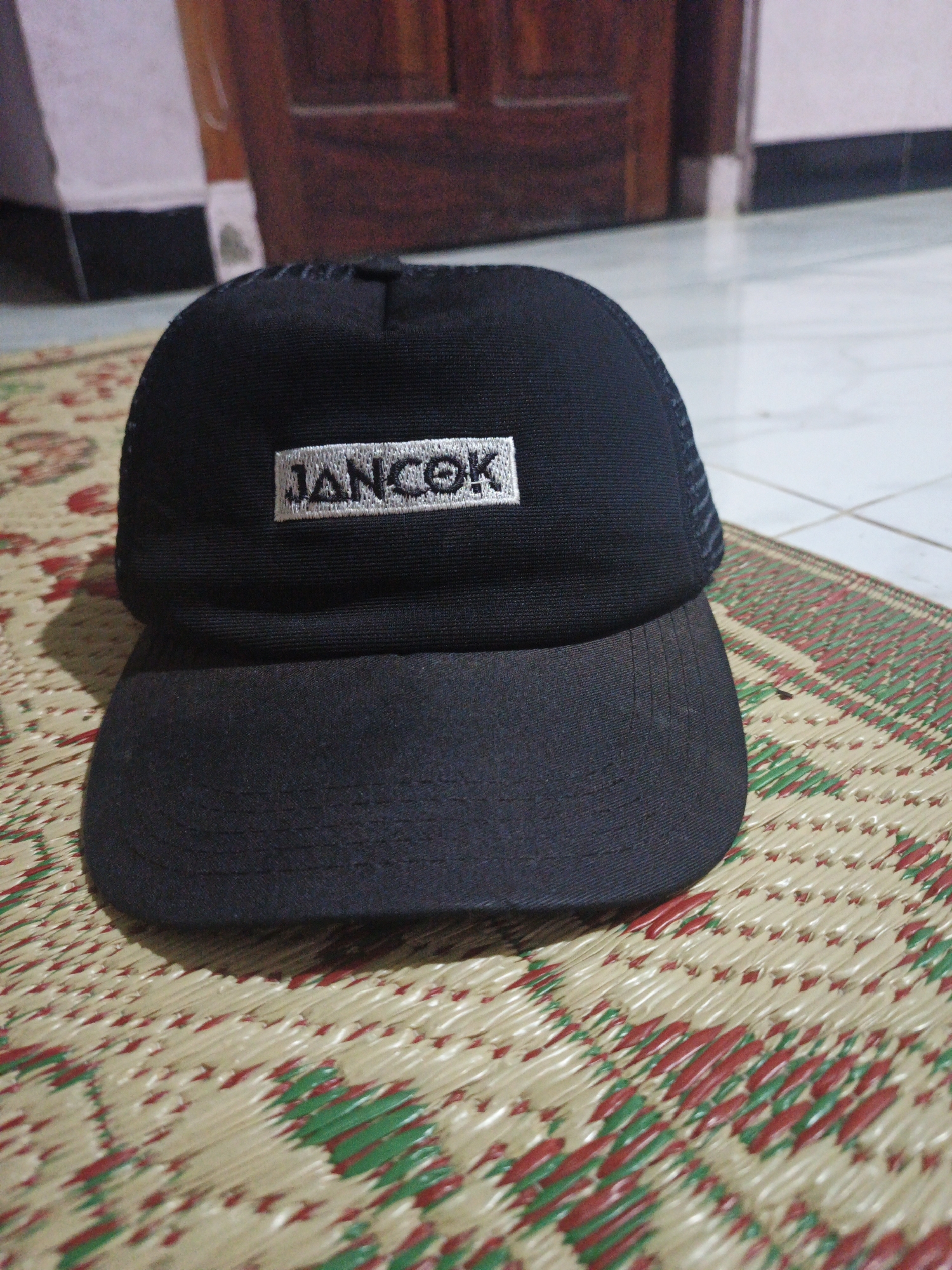 A black trucker hat with a white embroidered logo that says "JANCOK" is placed on a woven mat. The hat has a black mesh back and a black brim. The logo is centered on the front of the hat. The background shows a tiled floor and a wooden door. The scene is indoors, and no specific location in Kepel, Indonesia can be identified.