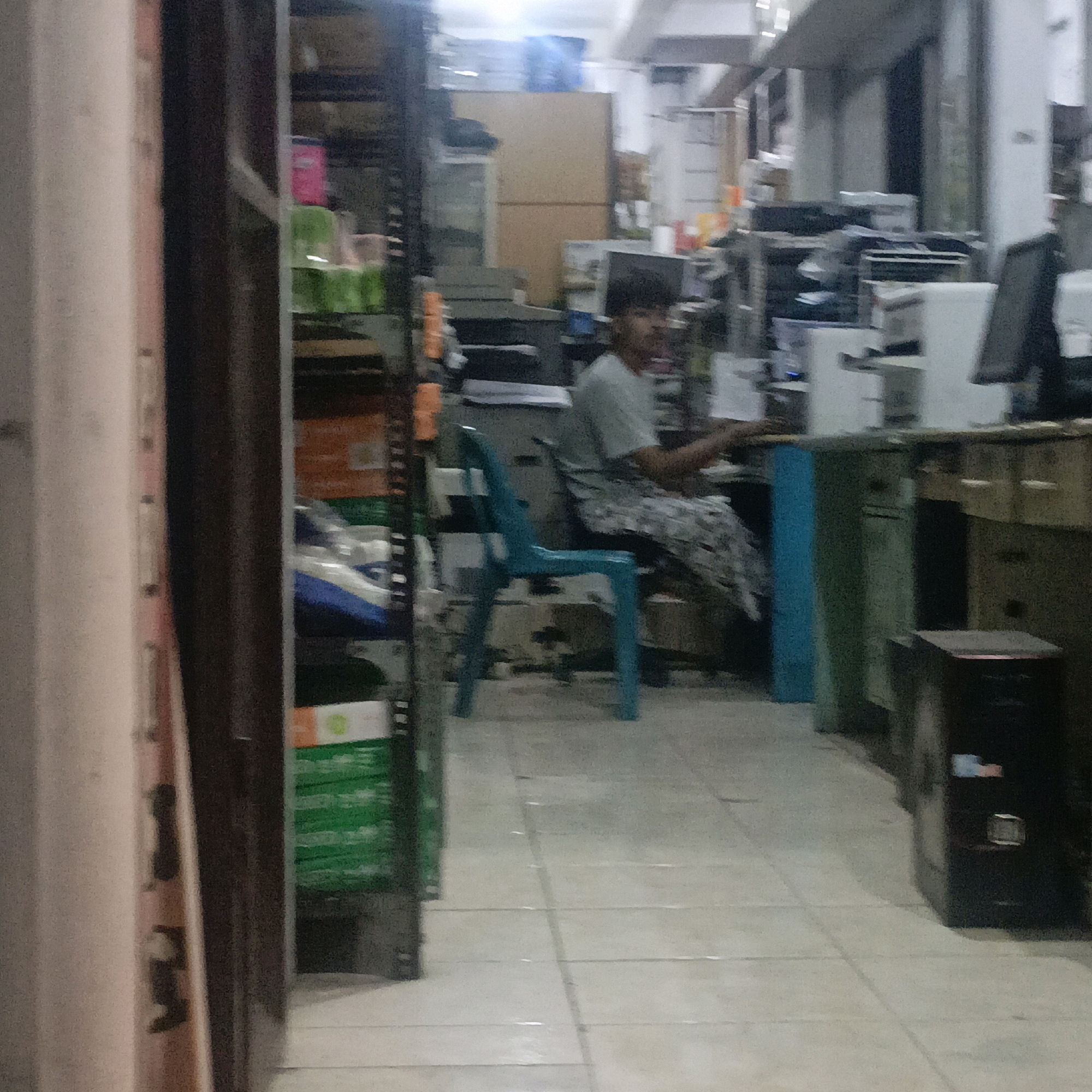 The image shows a man sitting in a blue plastic chair in what appears to be a store or office. He is wearing a grey t-shirt and patterned pants. The man is facing towards the right of the frame, with his hands resting on his lap. In the background, there are shelves stocked with various items, including what looks like boxes and containers. There is also a desk with office equipment, such as computers. The floor is tiled. The scene is located in Salatiga, Indonesia.