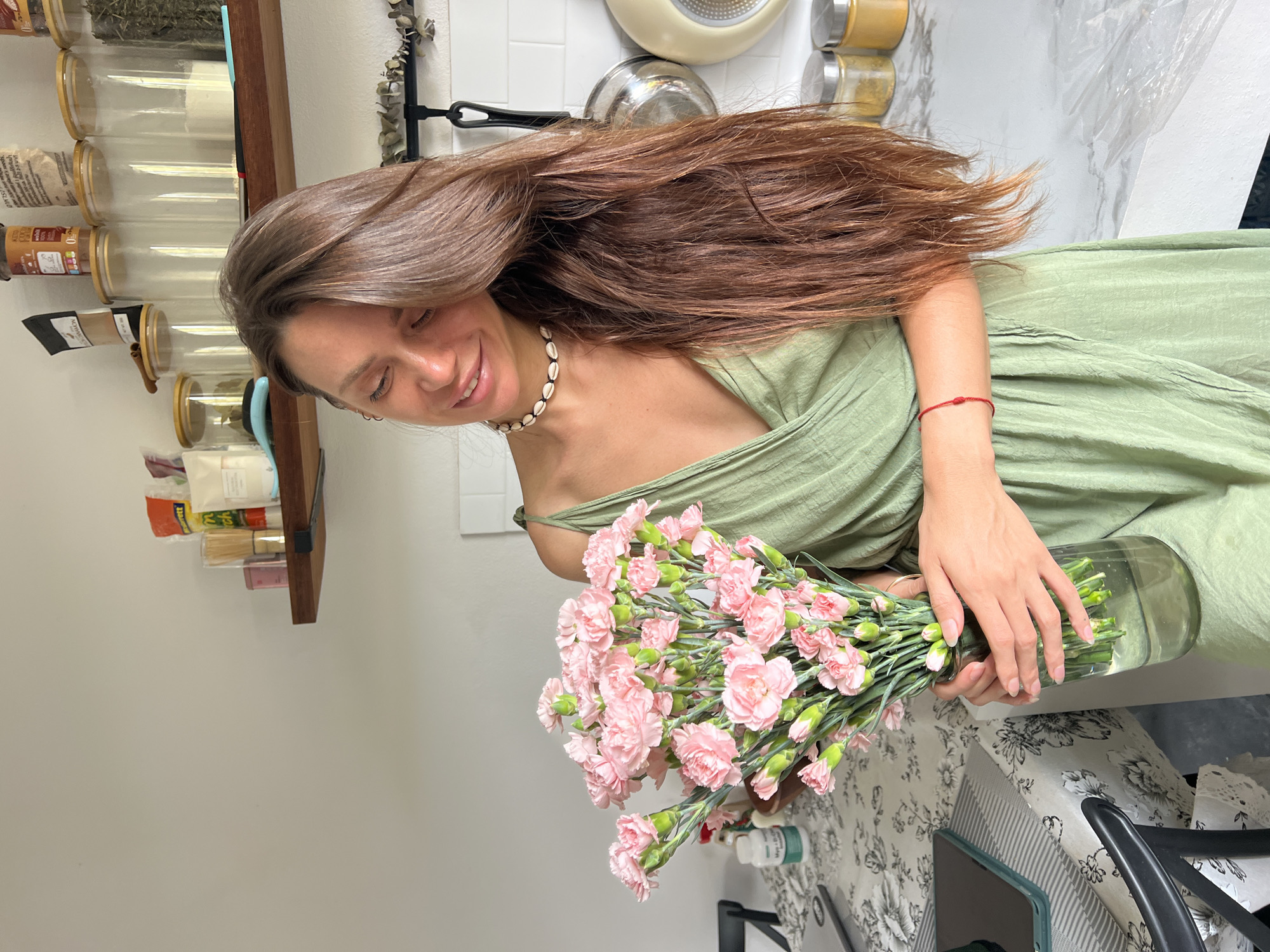 A woman with long brown hair, wearing a green dress and a shell necklace, is holding a vase filled with pink carnations. She is looking down at the flowers with a smile. The scene appears to be indoors, likely in a home kitchen, evident from the jars of spices on a shelf and cookware in the background. The table below is covered with a floral print cloth. The exact location is บ้านบางรักษ์, Thailand.