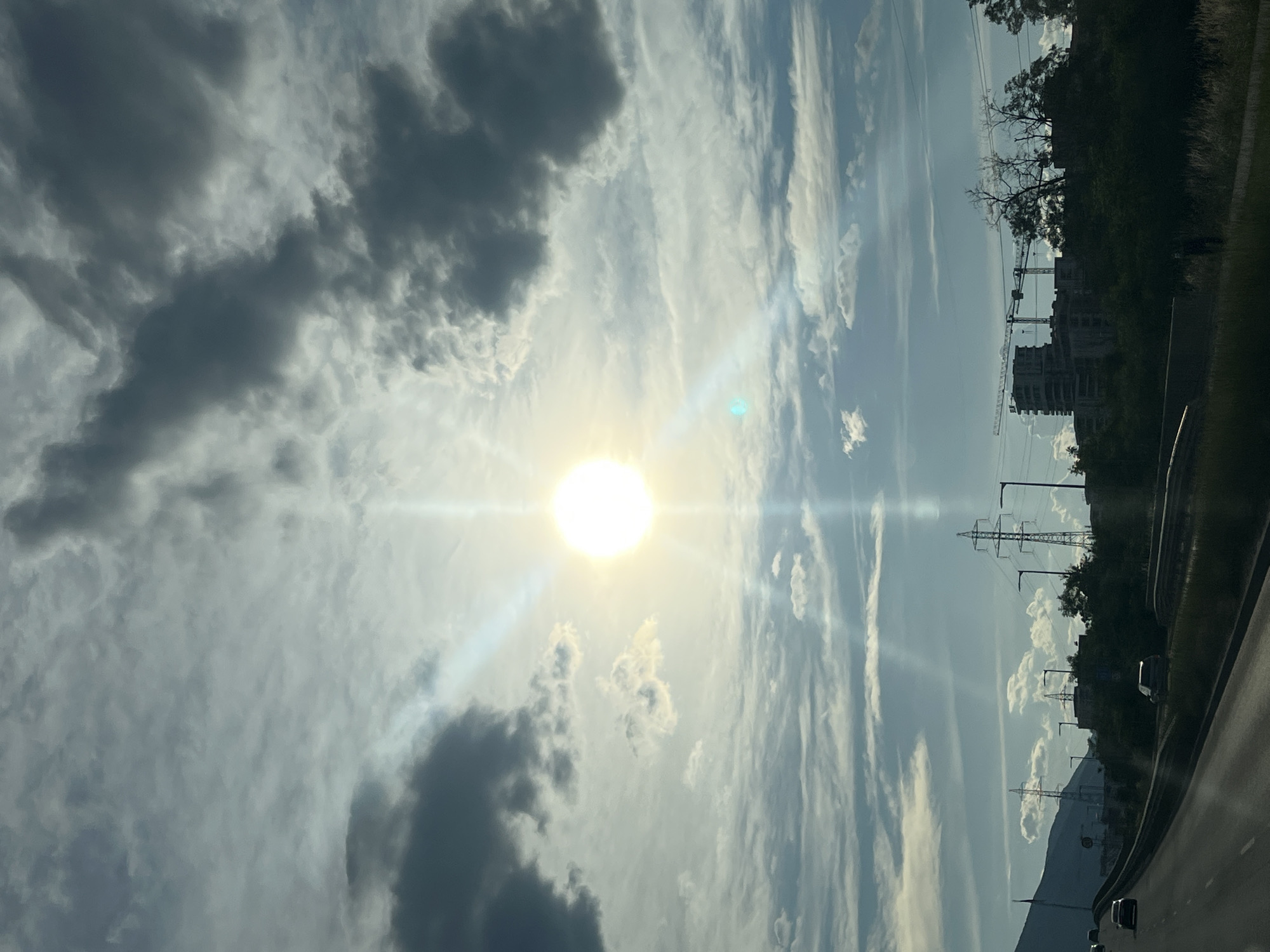 The image shows a sky with clouds and the sun shining brightly, creating a lens flare. There are also power lines, construction cranes, and buildings visible in the background, suggesting an urban or semi-urban environment. The presence of clouds indicates it might be daytime. The image is oriented vertically, so the scene appears to be viewed from a moving vehicle, likely on a road given the perspective of the foreground. The overall mood is bright and somewhat industrial. There is no visible text in the image.