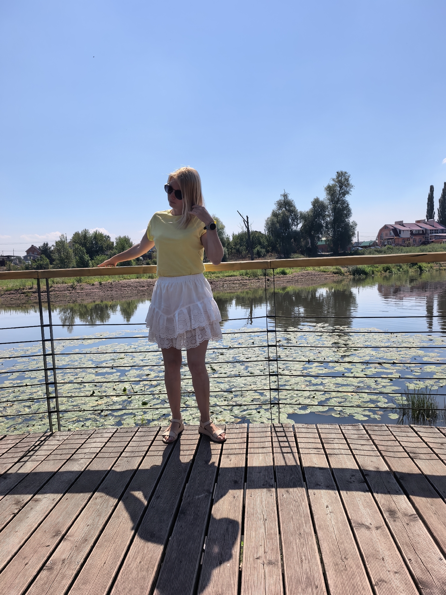 A Caucasian woman with blonde hair stands on a wooden pier, facing the water. She is wearing a yellow t-shirt and a white, tiered skirt. She has on sunglasses and sandals. Her right hand is raised, touching her hair. She is looking to her left. The pier overlooks a body of water filled with lily pads. Behind the water, trees and houses are visible under a clear blue sky. This scene is located in Sterlitamak, Russia.