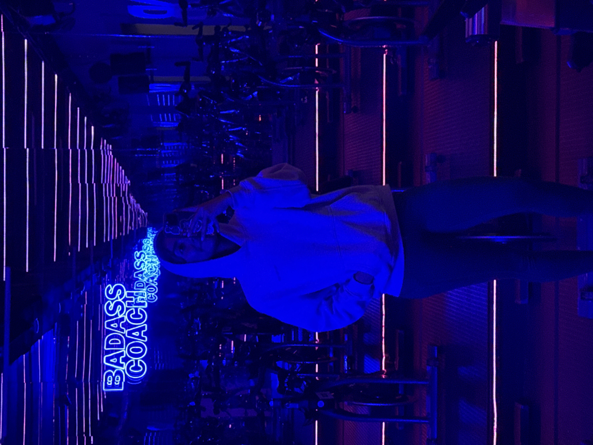 A photo taken indoors, rotated ninety degrees counter-clockwise, shows a woman in a hooded white sweatshirt and dark pants standing in a mirrored room with exercise bikes. Neon lights adorn the walls and ceiling, with "BADASS COACH" vertically displayed on one side. The room is bathed in blue and purple light, creating a nightclub-like atmosphere. The location is Puebla, Mexico.