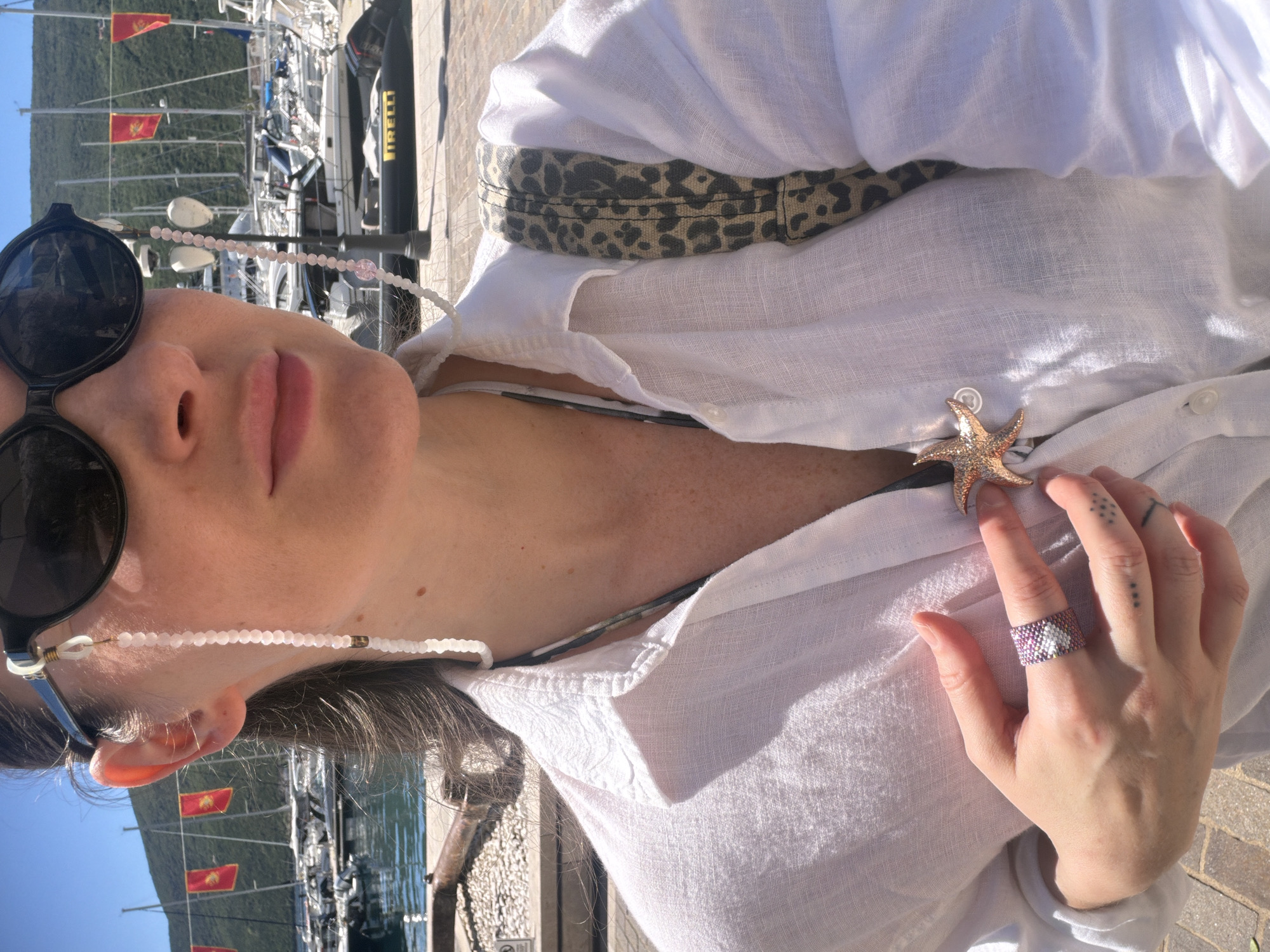 A medium, outdoor shot depicts a woman from a tilted perspective, likely at a marina in Đenovići, Montenegro. The woman wears sunglasses with a beaded chain, a white linen shirt adorned with a starfish brooch, and a ring crafted from pink and purple beads. She is partially visible, and the foreground features a stone-paved surface. In the background, boats are moored at the marina with flags of Montenegro waving. The day appears bright and sunny, evidenced by shadows and direct sunlight on the woman's skin.