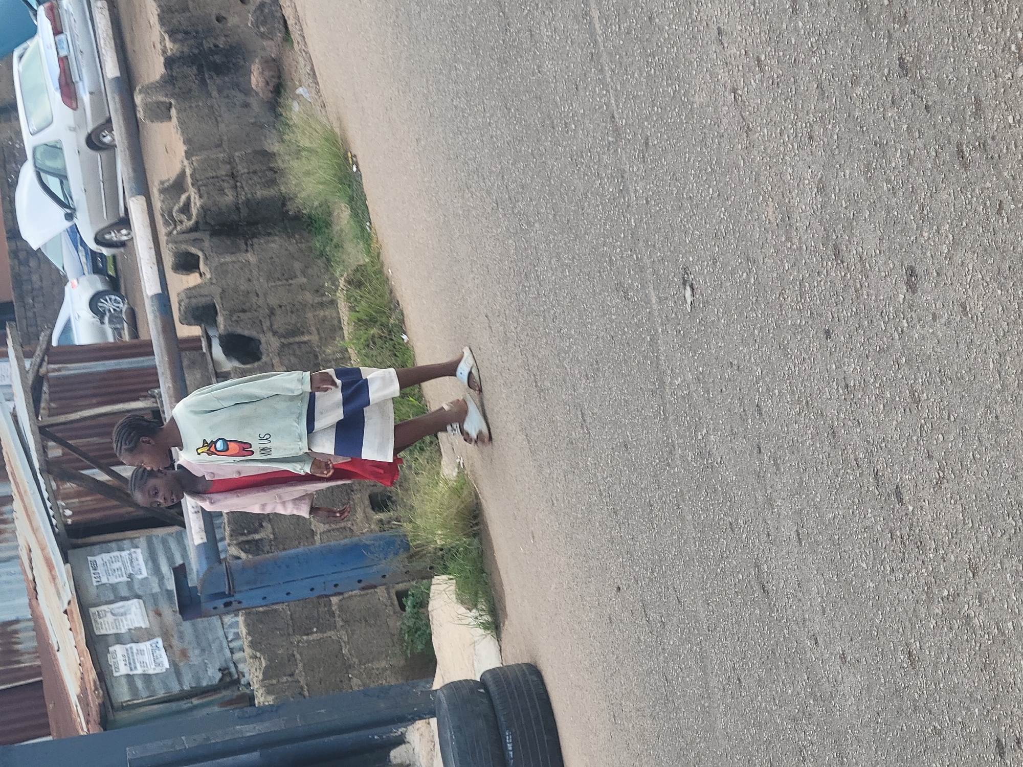 Two young females are walking on a paved road in Jos, Nigeria. The female on the left is wearing a pink coat over a red dress. The female on the right is wearing a light blue long-sleeved shirt with a graphic of a character from the game Among Us and blue and white striped shorts. Both are wearing sandals. Behind them, a brick wall separates the road from a building with a corrugated metal roof and some posters. Several cars are visible in the background, including a silver sedan with its hood up. Stacked tires are on the left side of the road. The environment appears to be an urban or semi-urban setting.