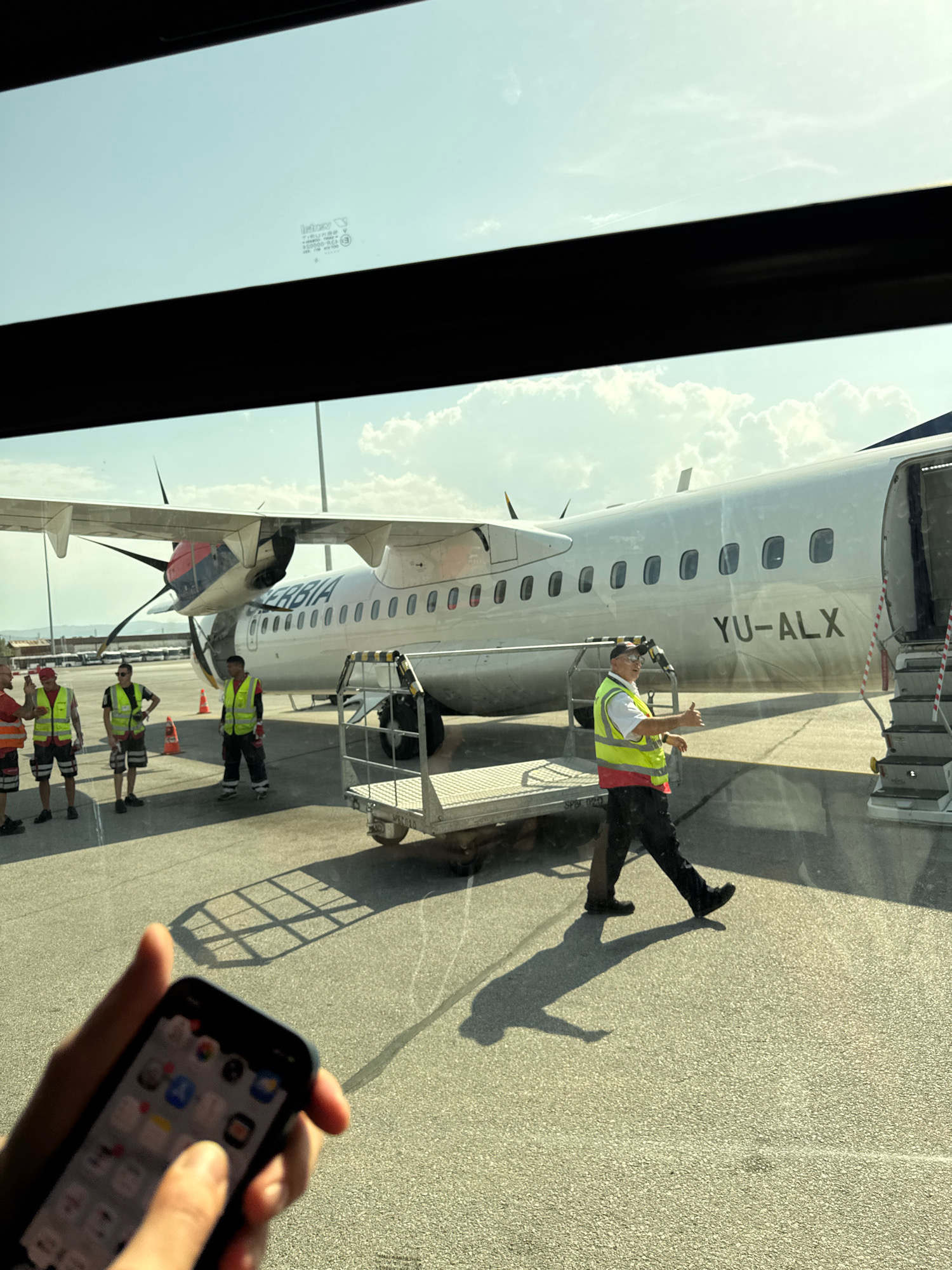 The image depicts an airport tarmac scene, viewed through a tinted window, likely from inside an airplane. In the foreground, a person's hand holds a smartphone displaying various app icons, with a person's thumb partially obscuring the screen. The background features a turboprop aircraft with "YU-ALX" painted on its fuselage and "SERBIA" above it. Several ground crew members in high-visibility vests are visible around the aircraft. One crew member, wearing a yellow vest and red undershirt, is walking towards the camera and giving a thumbs-up gesture. Another crew member is seen near a baggage cart. The scene is illuminated by bright sunlight, casting distinct shadows on the tarmac. The sky is clear with a few scattered clouds. This scene is identifiable as being at Sofia Airport (SOF) in Sofia, Bulgaria, due to the presence of JAT Airways (YU-ALX was a Serbian Airways aircraft) which historically operated flights to and from Sofia, and the typical appearance of airport infrastructure at SOF.