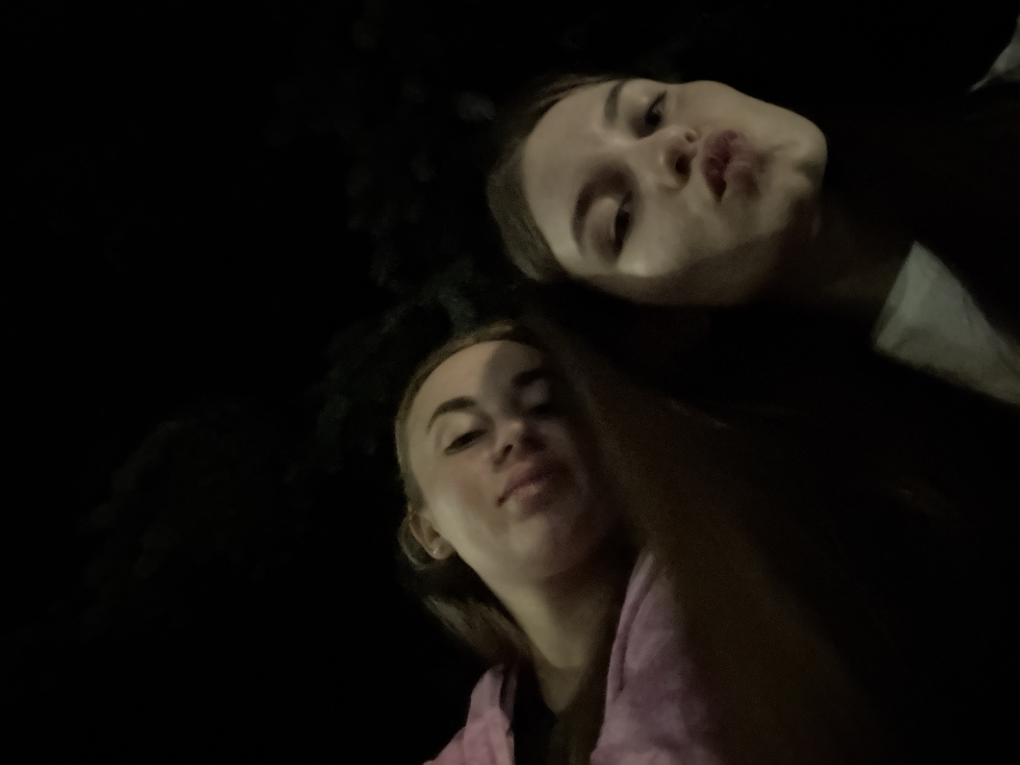 Two young women are captured in a low-angle selfie at night. The woman in the foreground, closer to the camera, has blonde hair and is wearing a pink hoodie. She is looking slightly upwards and to the left, with a soft expression. Behind her and slightly to the right is another young woman with dark hair. She is looking directly at the camera and puckering her lips. Both women appear to be outdoors, as indicated by the dark, indistinct background that suggests trees or foliage against a night sky. The lighting is dim, characteristic of nighttime, and the focus is primarily on the faces of the two subjects. The overall mood is casual and playful, likely taken during a social gathering or outing. The location context provided is Dnipro, Ukraine, but there are no specific visual cues within the image to confirm this. There is no visible text in the image.