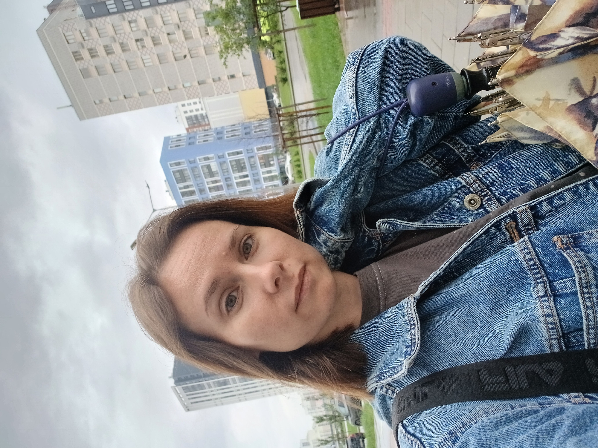 A medium shot selfie shows a woman with shoulder-length brown hair and light brown eyes, looking directly at the camera. She is wearing a denim jacket over a dark brown t-shirt. A black strap from a bag is visible over her left shoulder. A purple object, possibly an umbrella handle, is attached to a blue cord that hangs from her jacket. Part of a folded umbrella with a floral pattern is visible in the lower right corner.
The background depicts an overcast urban environment with tall, modern buildings. To the right, a grey building with a patterned facade rises several stories. To the left, a distinctive bright blue building with a grid-like window pattern is visible. The ground appears wet, suggesting recent rain. Trees and streetlights are also present. The buildings and architectural style are consistent with areas in Yekaterinburg, Russia, such as the Akademichesky district, known for its contemporary residential complexes.