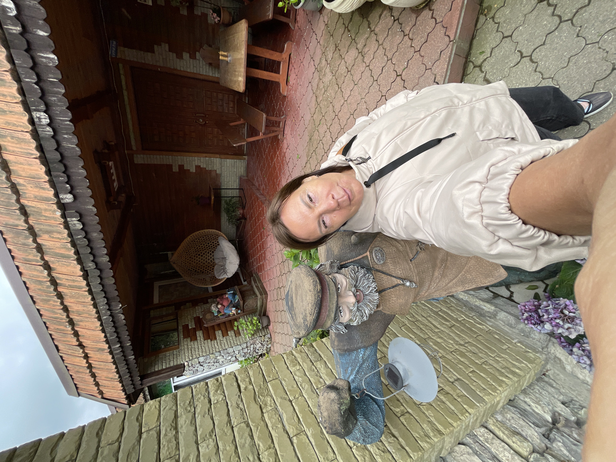 A woman is taking a selfie outdoors in Khartsyz Pervyi, Russia. She is wearing a light pink jacket, black pants, and dark shoes. Behind her is a statue of a man in a brown jacket, hat, and grey beard, standing on a yellow brick base with a solar-powered light. In the background, there is a house with a brown roof, wooden furniture, and hanging wicker chair. The ground is paved with grey stone.