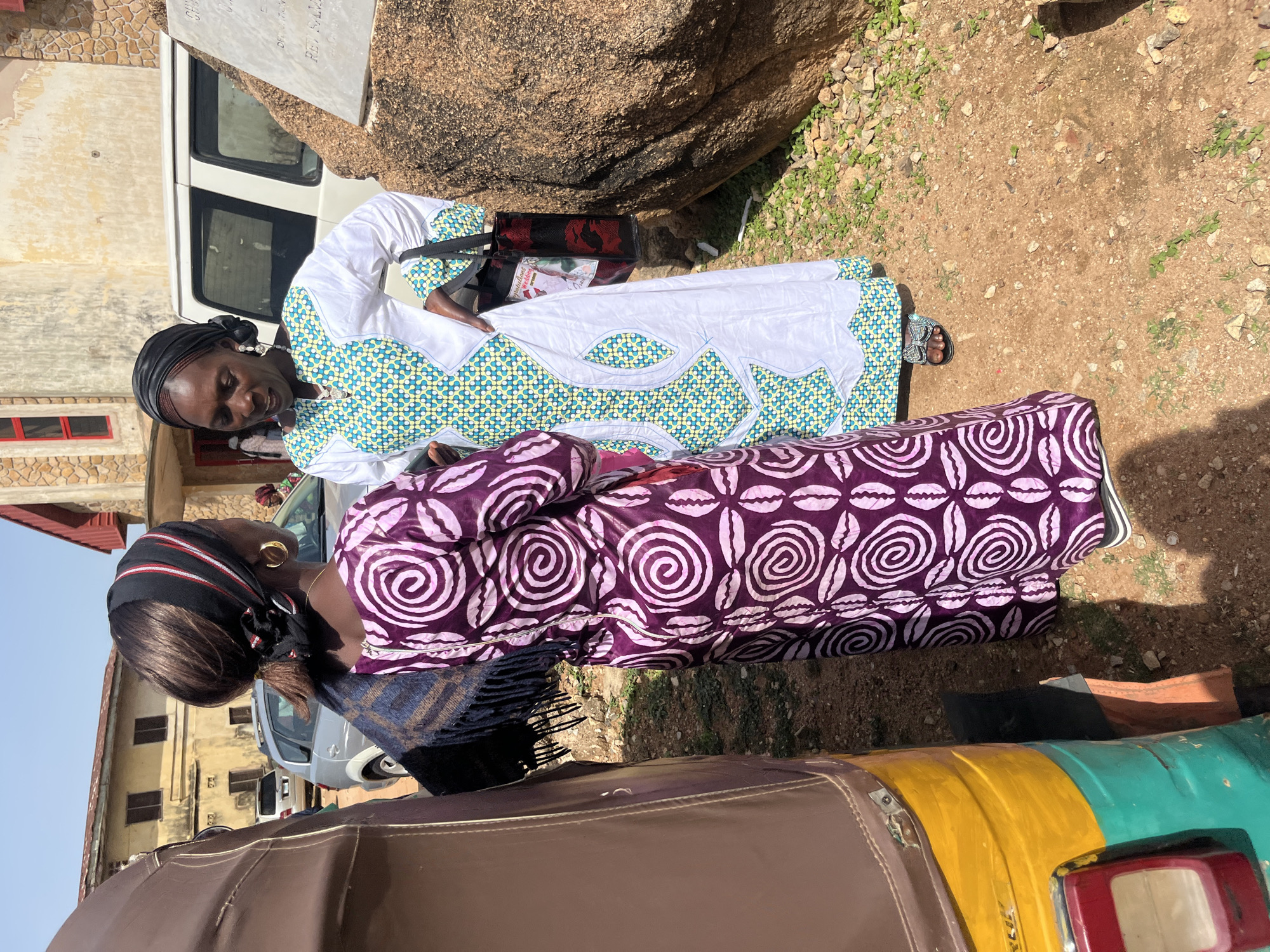Two women stand facing each other in Bukuru, Nigeria. The woman on the left, facing away from the camera, wears a purple and white patterned dress and a dark headscarf. She has a blue and brown patterned scarf draped over her shoulders. The woman on the right, facing the camera, wears a white dress with a teal and yellow geometric pattern on the bodice and sleeves. She has a black headscarf and large hoop earrings. A brown and yellow auto-rickshaw is partially visible on the left side of the frame. In the background, a building with yellow walls and a red-roofed section is visible, along with a white van and a large rock formation. The ground is covered in dirt and scattered stones.