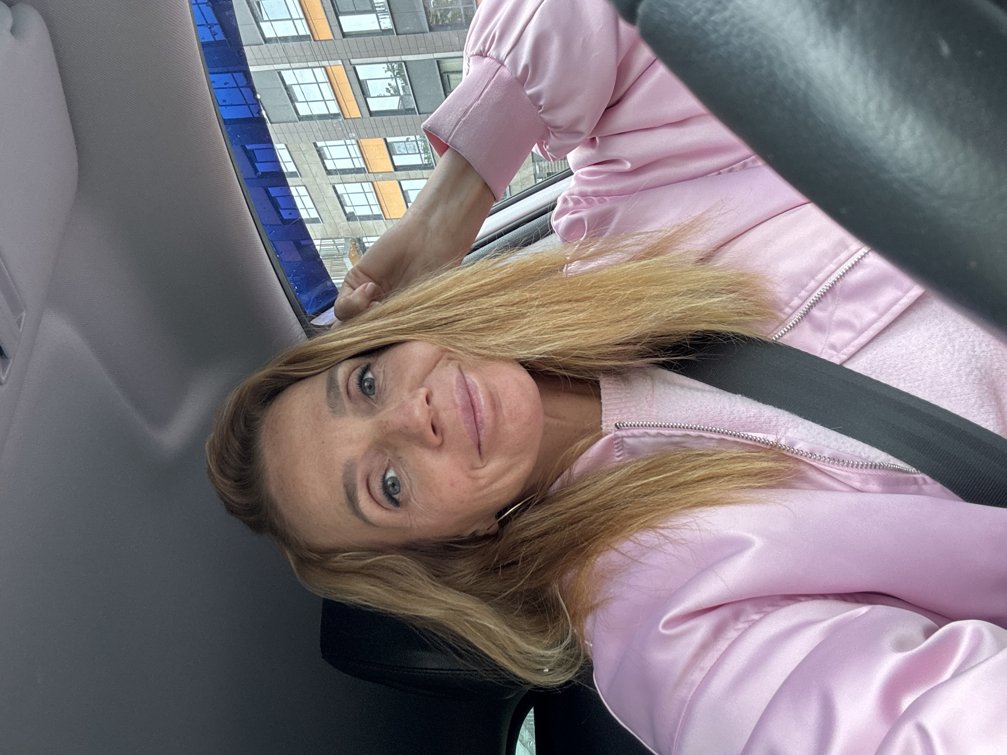 A woman is in a car, wearing a pink jacket and a seatbelt. The background shows a building with multiple windows, indicating an urban environment, consistent with Saint Petersburg, Russia. The image orientation is tilted.