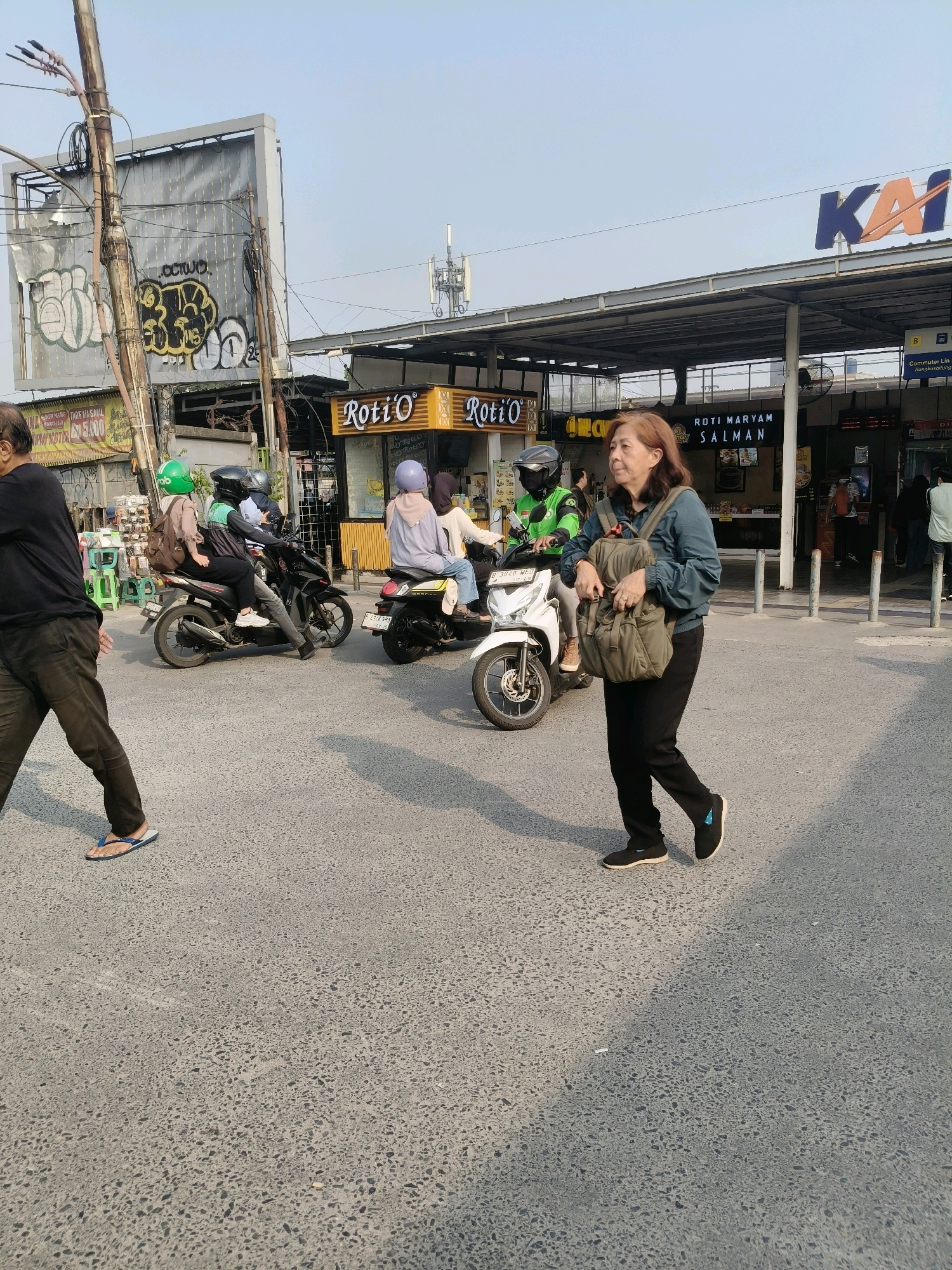 The image shows a street scene in South Tangerang, Indonesia, likely near a train station as indicated by the "KAI" logo in the background, which is the logo of the Indonesian state-owned railway company.
In the foreground, a woman with shoulder-length brown hair is walking towards the right of the frame. She is wearing a dark teal jacket, black pants, and black athletic shoes. She is carrying a light olive-green backpack slung over her left shoulder. To the left of the woman, a person in a black shirt and dark pants is walking away from the camera, wearing flip-flops.
The middle ground features several motorcycles on the road. One motorcycle, ridden by a person wearing a green helmet and a light brown jacket, has a passenger in a pinkish-purple top and a light brown headscarf. Another motorcycle, ridden by a person in a black helmet and a dark green jacket, has a passenger wearing a light purple top and a light brown headscarf. A white motorcycle is parked further back.
In the background, there are several stalls or kiosks. Two prominent signs read "Roti'O," indicating a bakery chain. Another sign above a stall reads "ROTI MARYAM SALMAN." Above these structures, a large billboard covered in graffiti is visible, along with utility poles and wires. In the far background, a cell tower and a building with the "KAI" logo can be seen. The environment appears to be an urban setting with a paved road. The lighting suggests it is daytime, with shadows cast on the road.